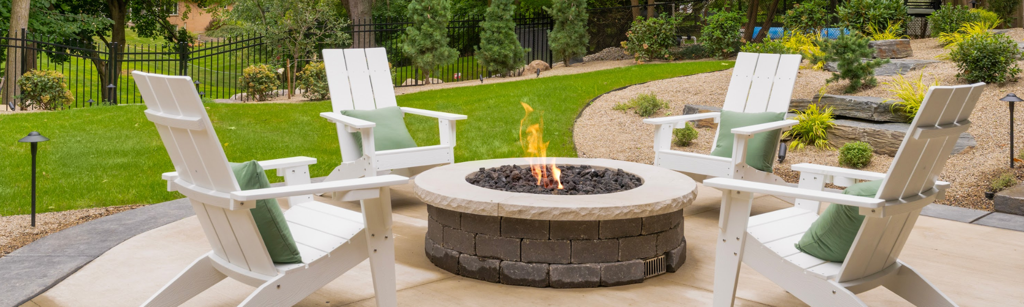 A backyard patio with four white Adirondack chairs around a round fire pit, with a landscaped yard and trees in the background.