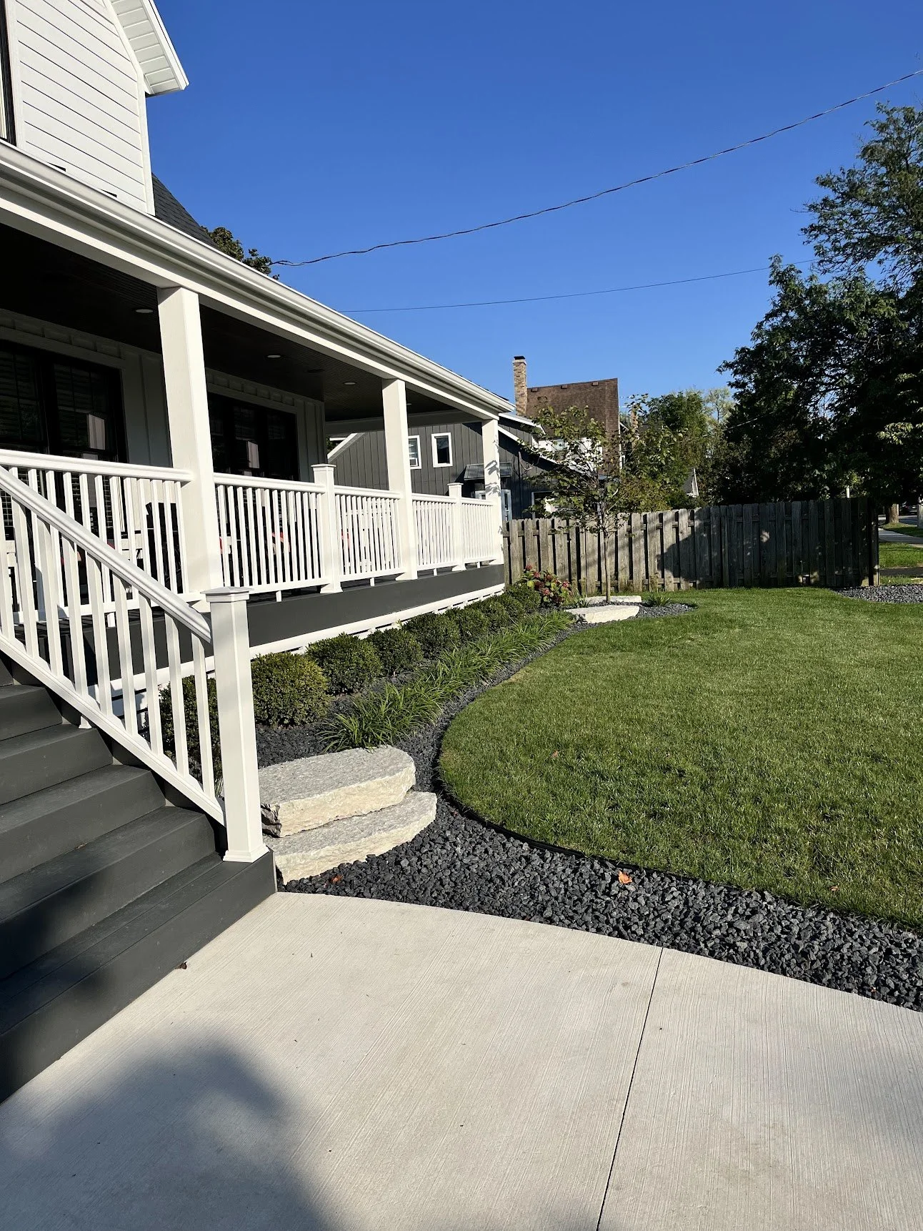 View of a house's backyard showing a deck with white railing, a green lawn, landscaped plants and rocks, and a wooden fence under a clear blue sky.