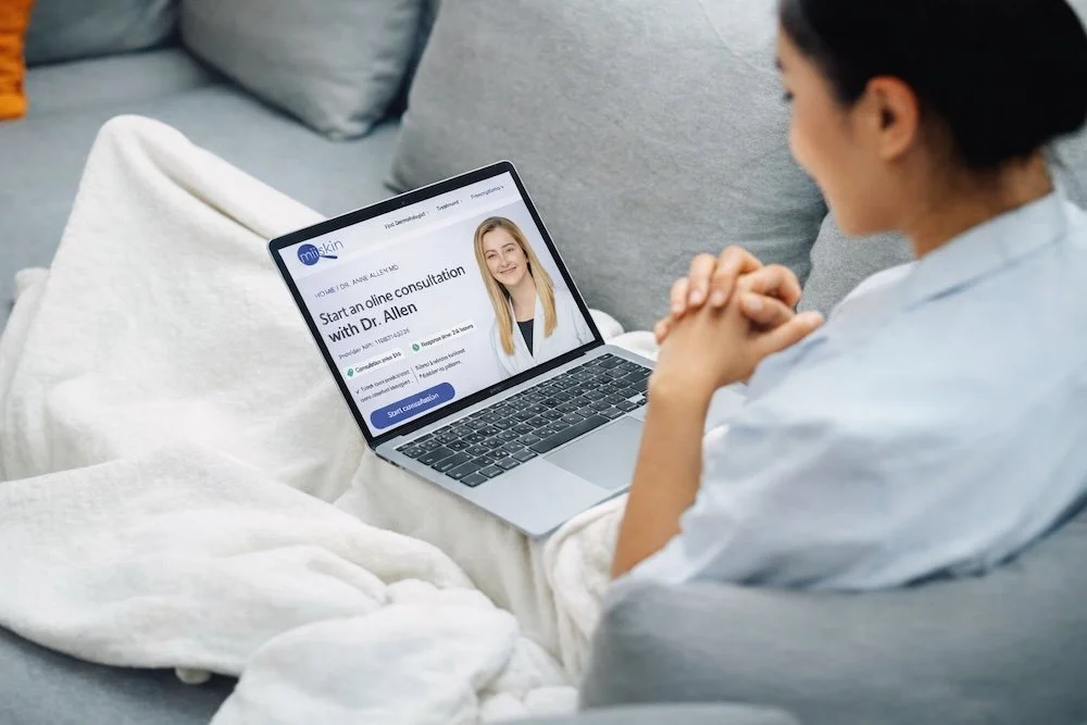 A woman sitting on a sofa using a laptop to start an online consultation with Anne Allen, dermatologist.