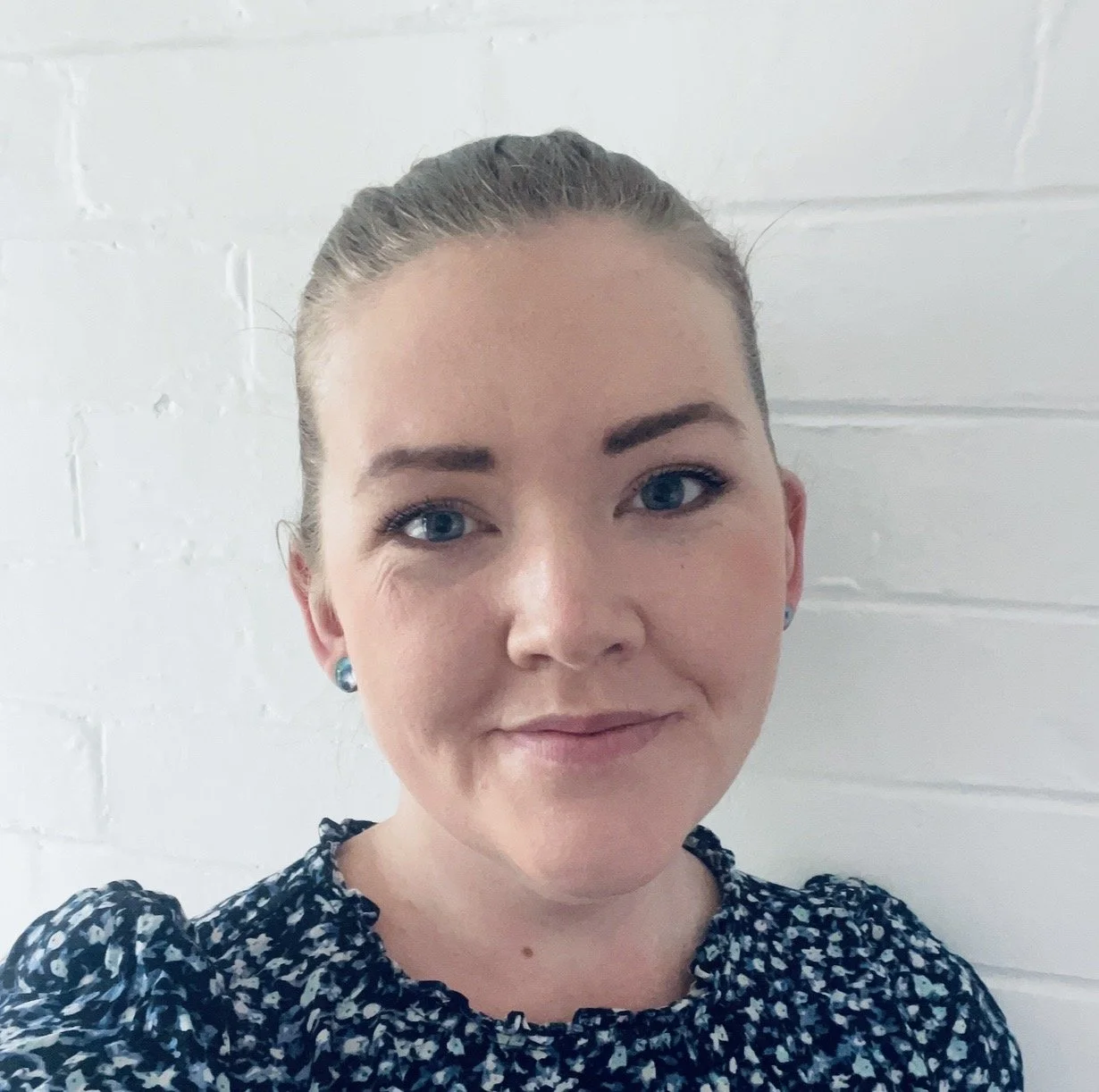 A young woman with light skin, blue eyes, and dark earrings taking a selfie in front of a white brick wall.
