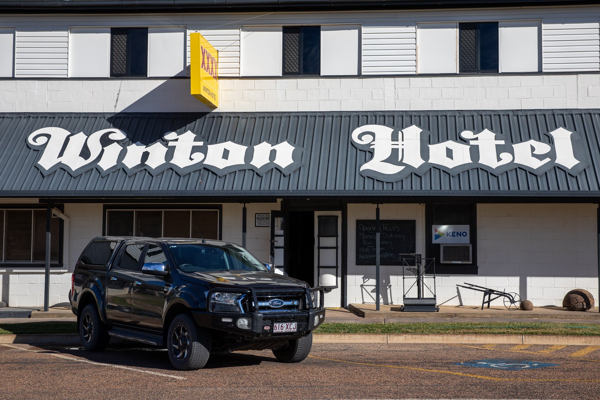 Front view of Watson Hotel with a black Ford pickup truck parked in front, large white signage on the dark gray building, and a sign indicating it is open on the black window to the right.