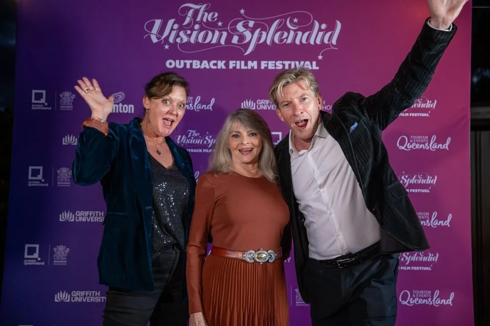 Three smiling people standing in front of a purple banner at The Vision Splendid Outback Film Festival, posing for a photo. The woman on the left is wearing a navy jacket and black shirt, raising her hand. The woman in the middle is wearing a rust-colored dress with a wide belt. The man on the right is wearing a black jacket, white shirt, and has his arm raised in excitement.