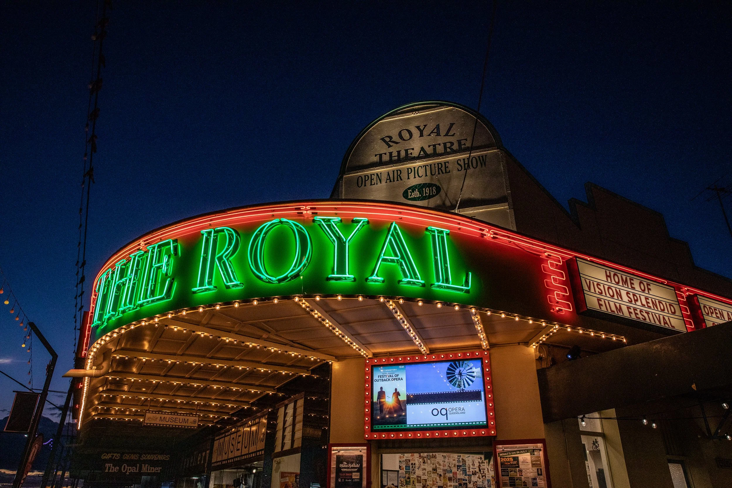 The Royal Theatre marquee illuminated with green and red neon lights at dusk, featuring a digital screen displaying advertisements and festival information.