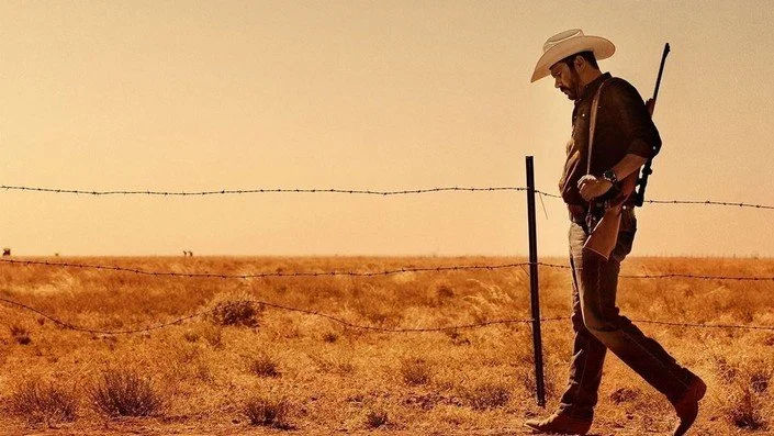 A man wearing a cowboy hat, sunglasses, and dark clothing walking through a dry, grassy desert with a rifle slung over his shoulder, near a barbed wire fence at sunset.
