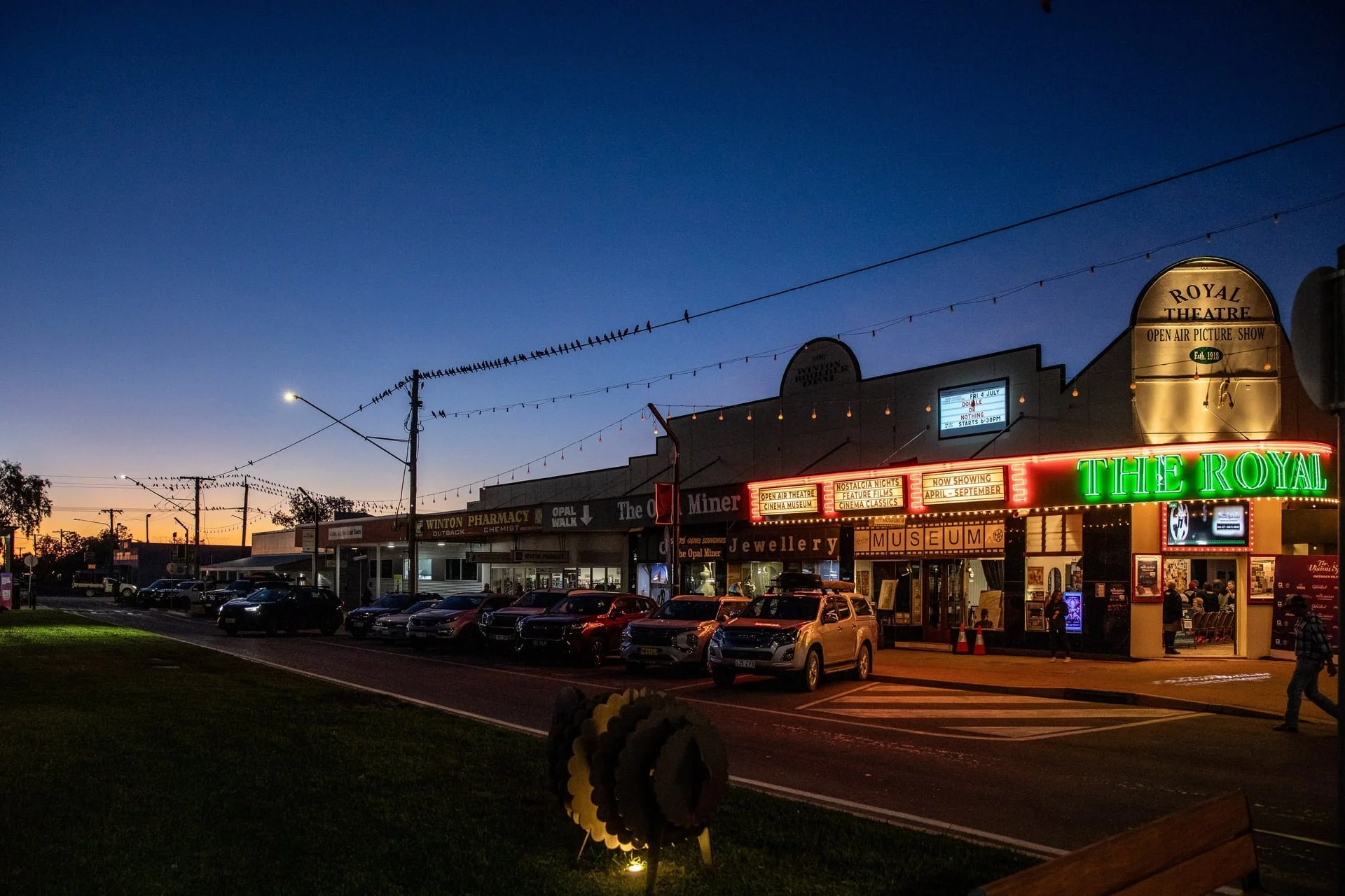 Nighttime view of the Royal Theatre with neon lights and marquee signs, parked cars along the street, and a pedestrian crossing.