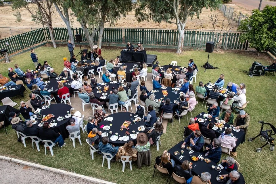 Breakfast gathering with multiple round banquet tables covered with black tablecloths, surrounded by white and colored chairs, with people eating and socializing, set on a grassy area with trees, a fence, and sound equipment in the background.
