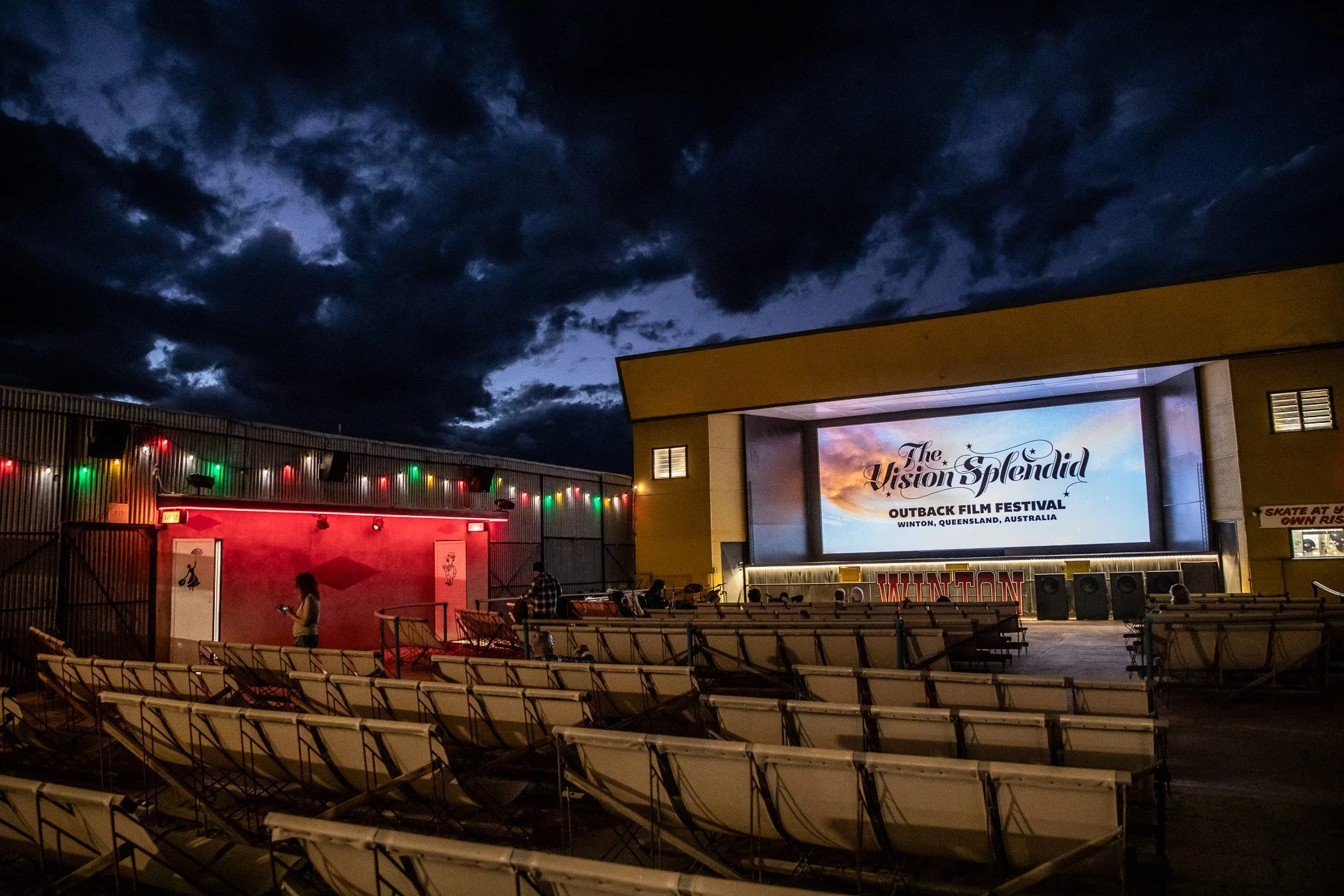 An outdoor outdoor movie theater with a large screen displaying 'The Vision Splendid Outback Film Festival Winton, Queensland, Australia' at dusk. Empty chairs are arranged in front of the screen, and a few people are standing near a wall with colorful lights under a dark, cloudy sky.