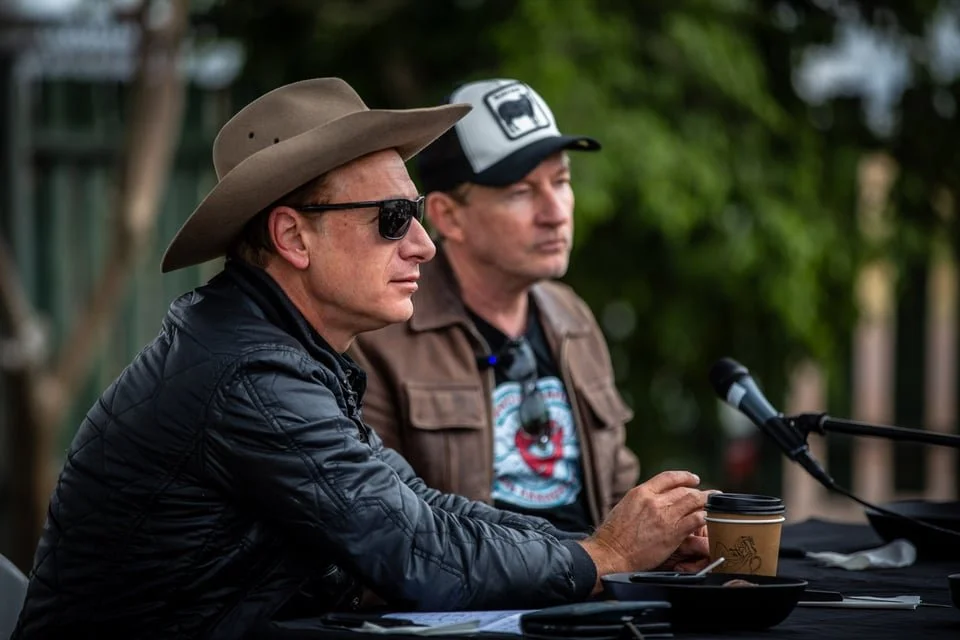 Two men sit at a table outdoors, one wearing a large cowboy hat, sunglasses, and a black jacket, the other wearing a baseball cap, a brown jacket, and a graphic T-shirt. There is a microphone and a coffee cup on the table.