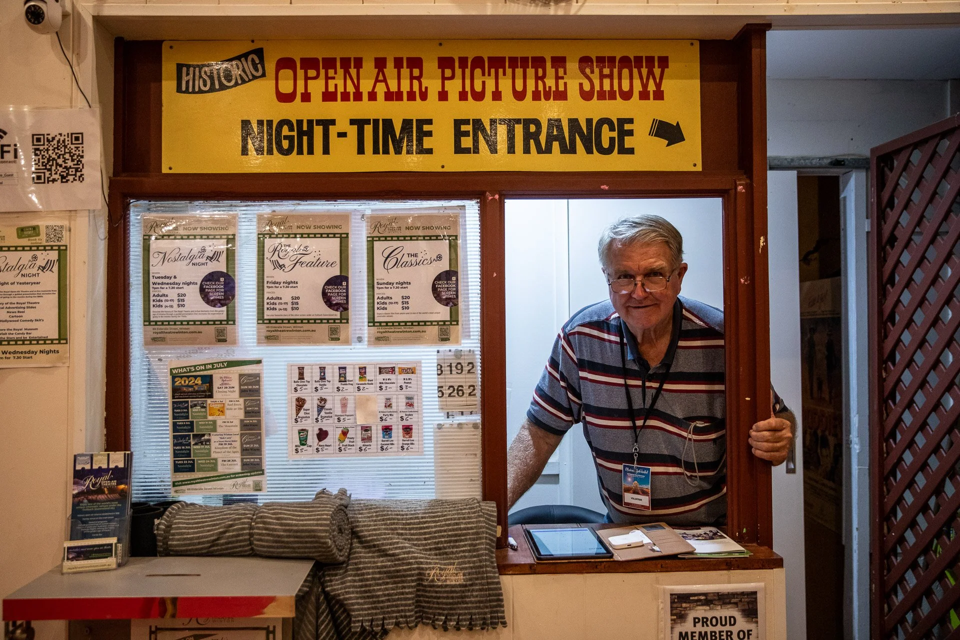 A man with glasses and gray hair smiling and leaning out of a ticket booth window, behind which is a sign that reads "OPEN AIR PICTURE SHOW NIGHT-TIME ENTRANCE" with an arrow. The booth has posters advertising movies and a table with a tablet, clipbo