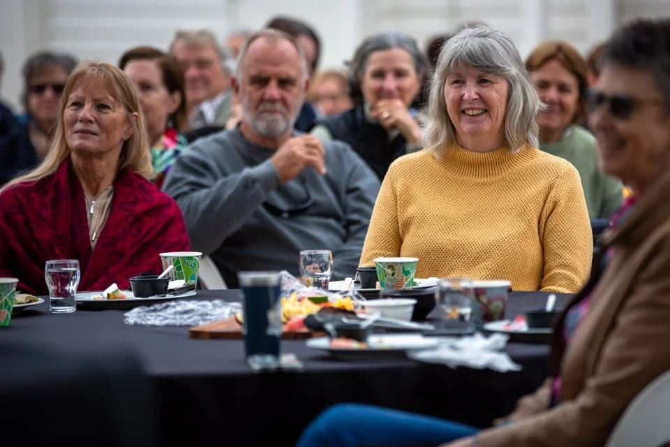 A group of people sitting at a table, smiling and engaging in a social gathering, with food and drinks on the table at the Vision Splendid Film Festival.