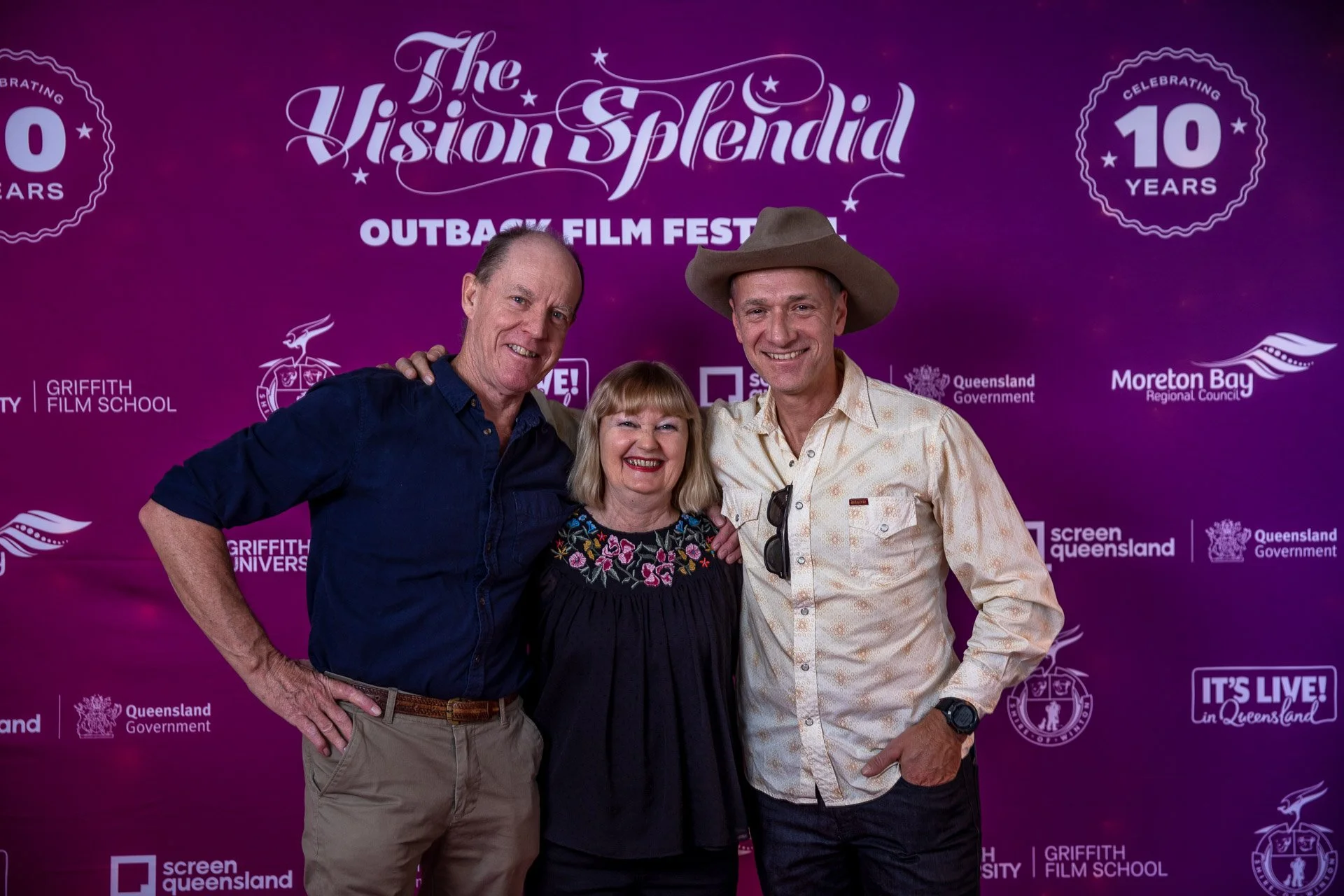 Three individuals smiling and posing together at the Outback Film Festival, standing in front of a purple backdrop with event logos and text.