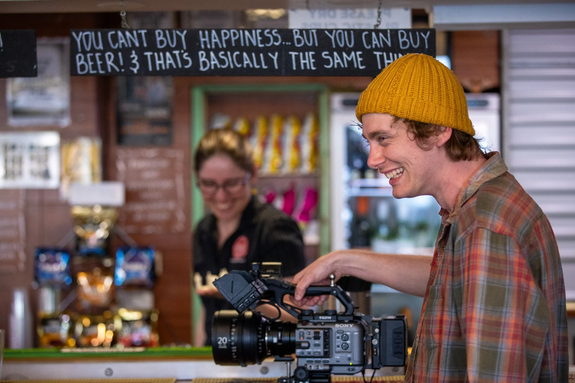 A man in a yellow knit cap and plaid shirt filming with a professional camera at a bar counter, with a smiling woman in black behind the counter and a sign about beer in the background.