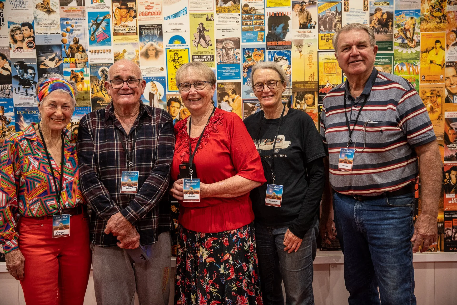Five older volunteers standing together in front of a colourful collage of vintage movie posters, all smiling at the camera, wearing casual clothes and event badges.