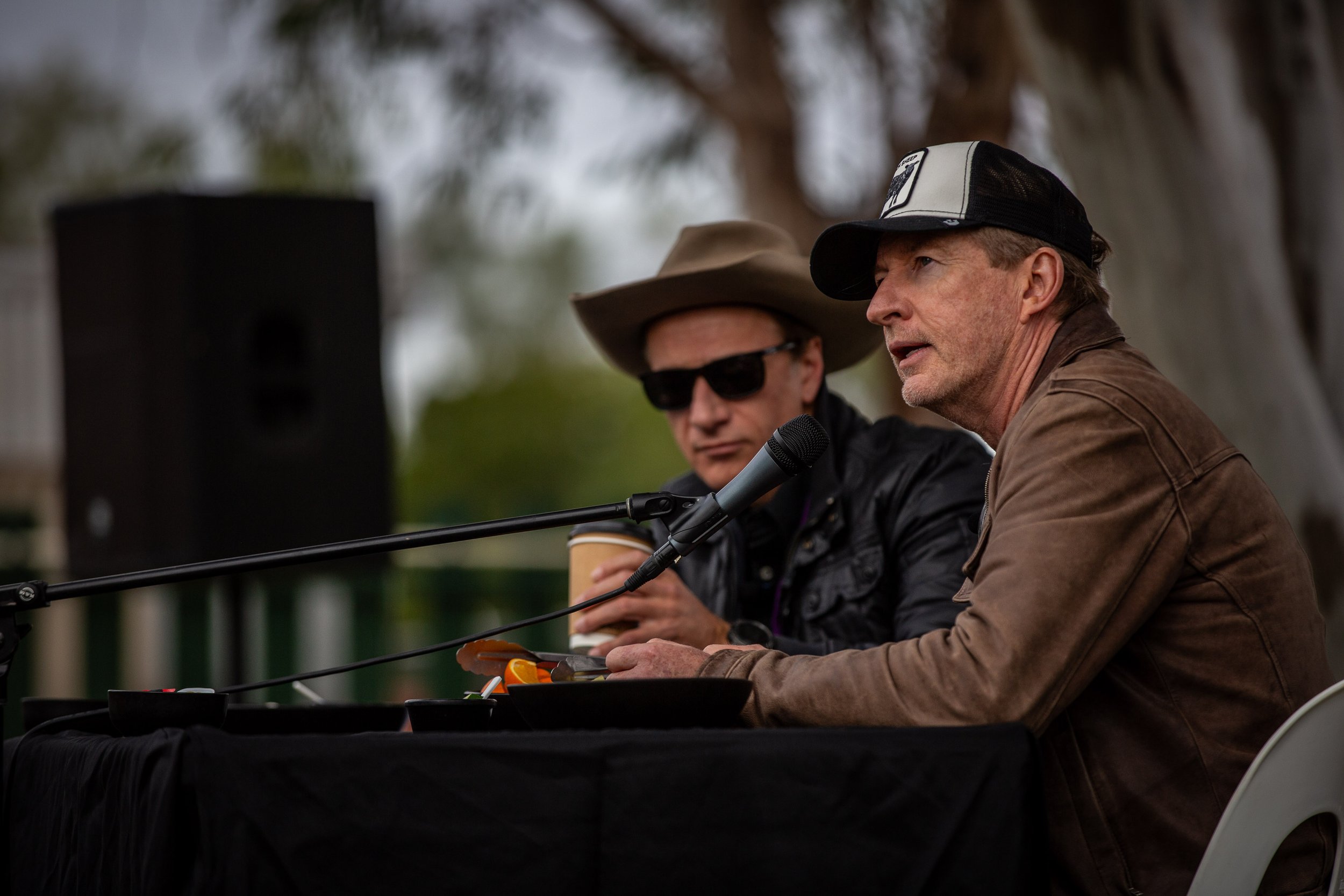 Two men sit at a table outdoors, speaking into a microphone. One wears a brown leather jacket and a black and white cap, the other wears sunglasses, a hat, and a jacket. A speaker is in the background.