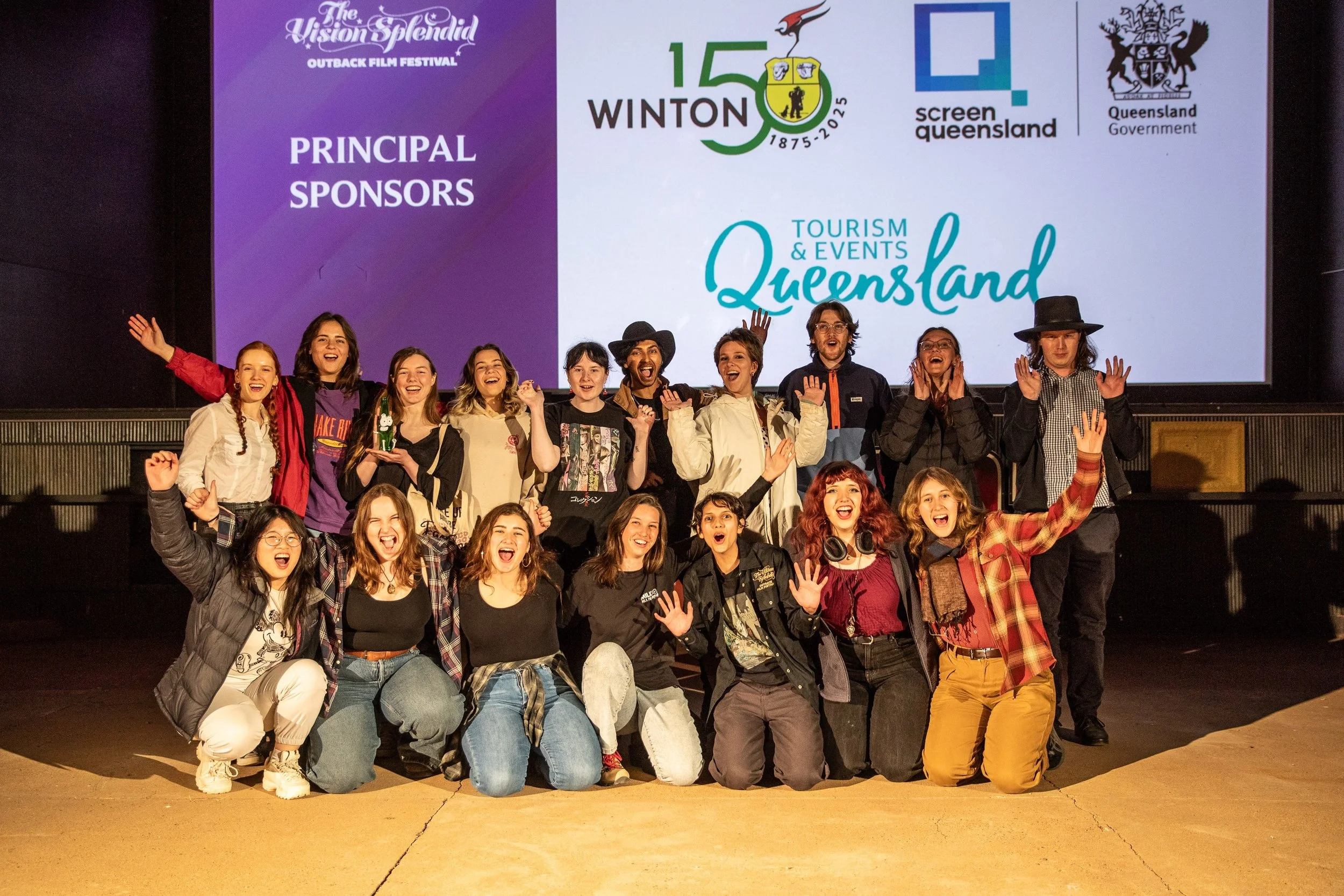 Group of young people on stage at the Outback Film Festival, celebrating with smiles and raised hands, in front of a screen displaying logos and text for Winton, Queensland, and Tourism & Events Queensland.