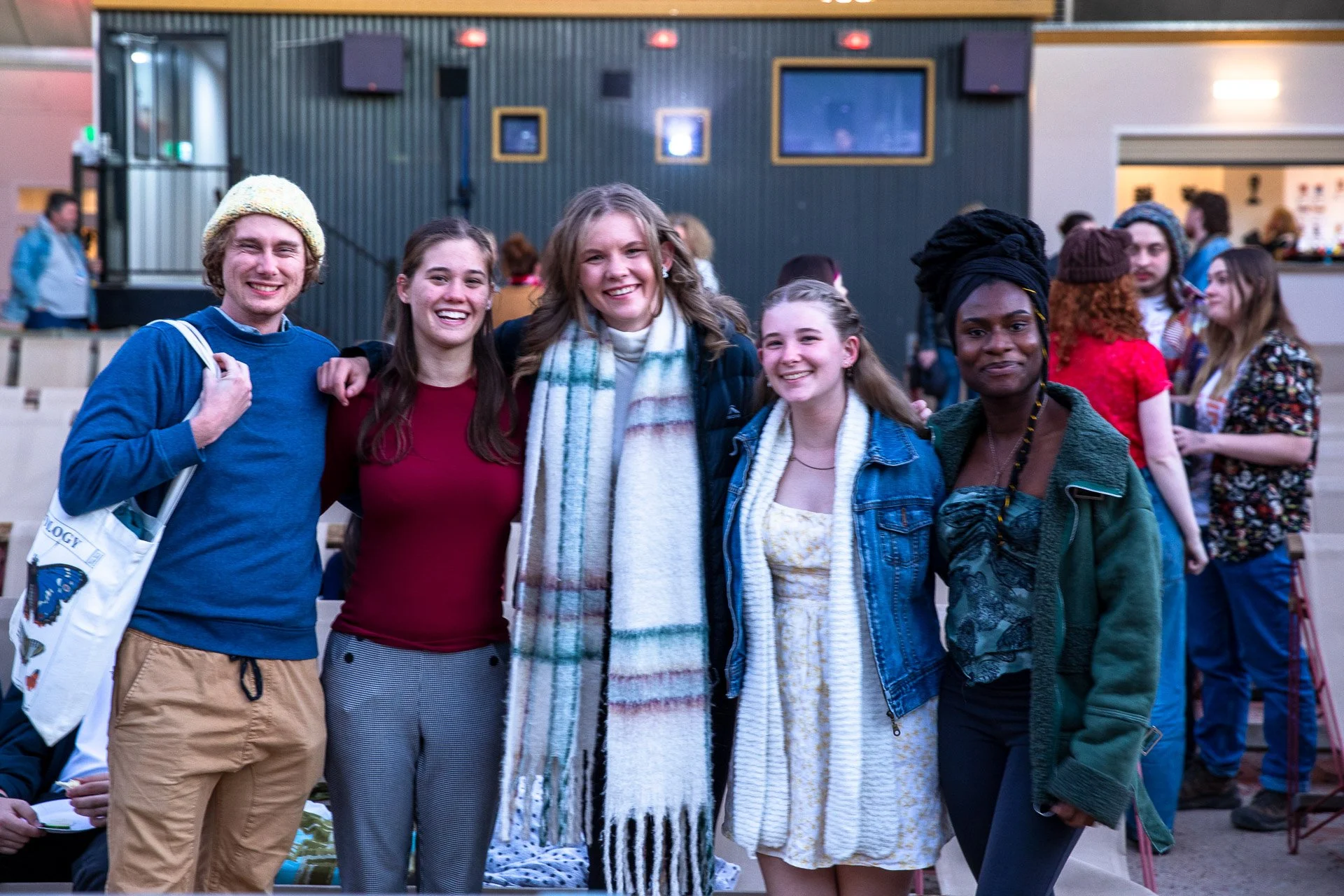 Group of five smiling young people standing together indoors at a social event, with other attendees and a candy bar kiosk in the background.