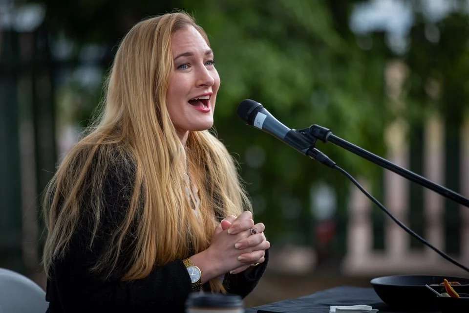 A woman with long blonde hair speaking into a microphone outdoors, smiling, with her hands clasped together.