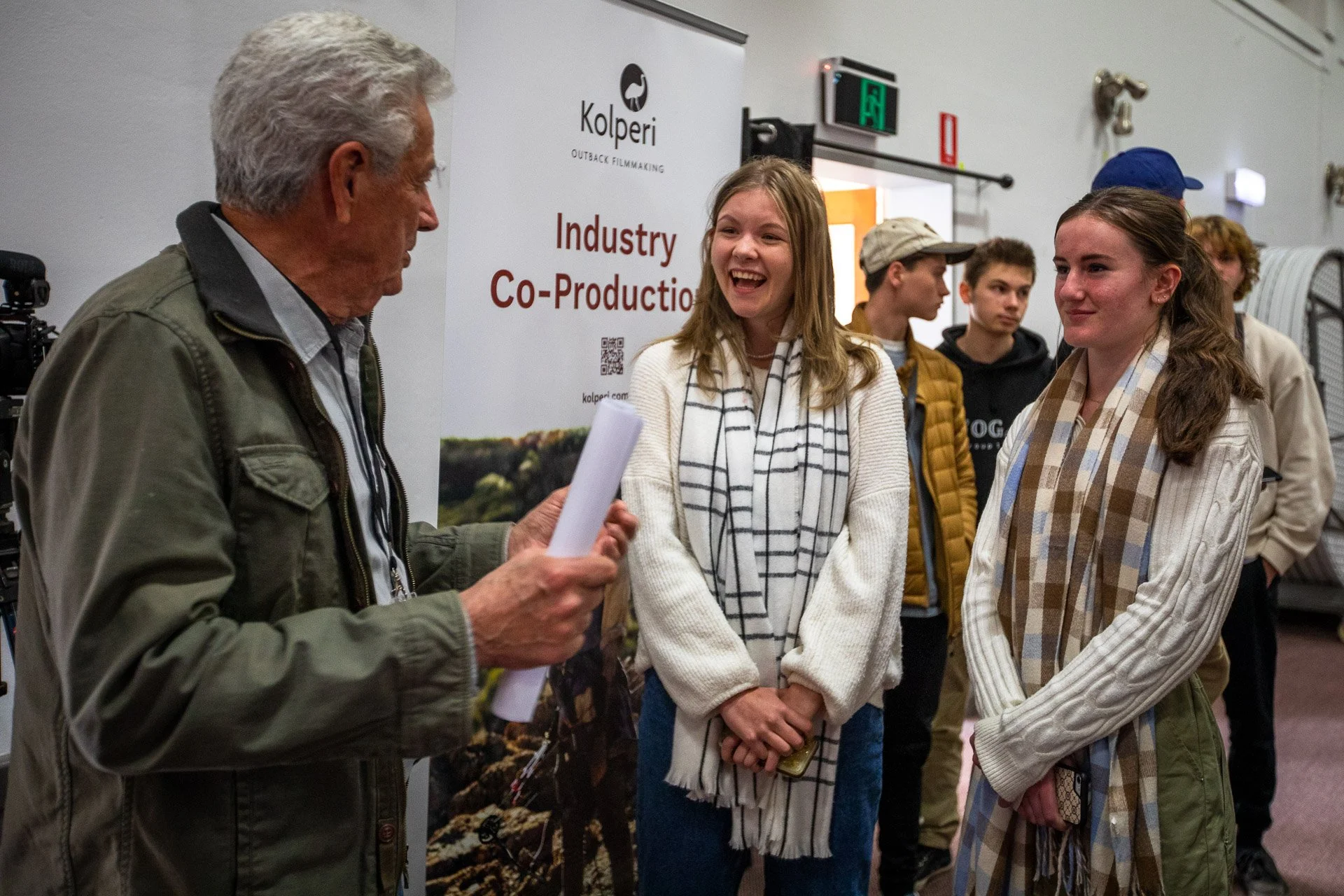 An older man with gray hair, wearing a green jacket, is talking to two young women, one with blonde hair and another with brown hair, at an indoor event. The background has a banner with the words 'Kolperi Industry Co-Production' and a small group of young people, some wearing jackets, look on.