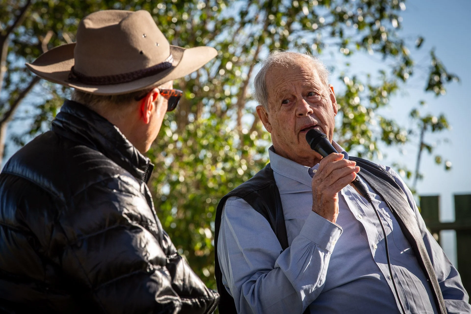 Two elderly men outdoors, one speaking into a microphone, the other listening, with trees and blue sky in the background.