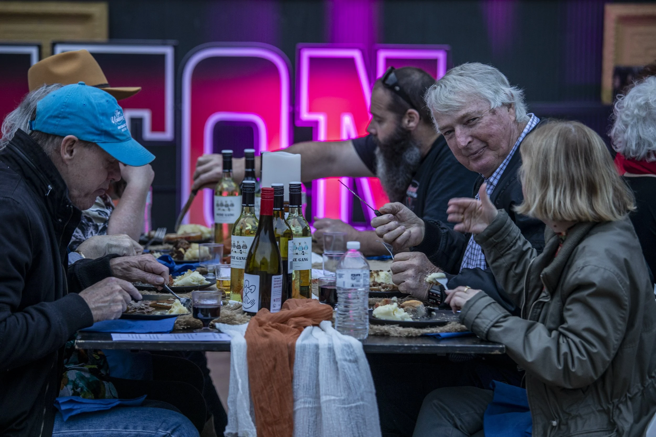 People enjoying a meal at a long table with bottles of wine and food, in a dimly lit setting with neon lights in the background.