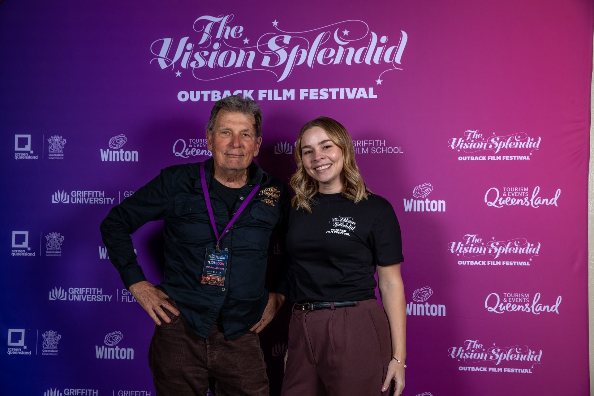 Two people standing in front of a purple backdrop at the Outback Film Festival. The man on the left is wearing a black shirt and has a purple lanyard. The young woman on the right is smiling, wearing a black t-shirt with the festival logo and maroon pants.