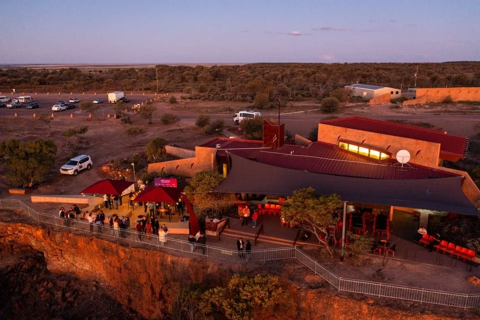 An evening outdoor scene at the Australian Age of Dinosaurs museum with many people, some seated under canopies, on a patio above a cliff, with a parking lot and outback landscape in the background.