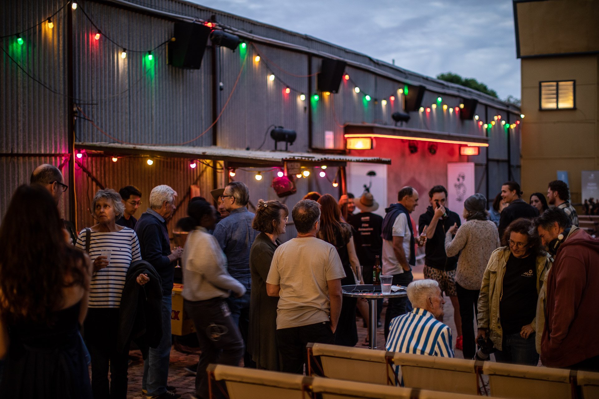 People socializing at an outdoor theatre gathering during early evening, with colorful string lights hanging on a corrugated metal building.