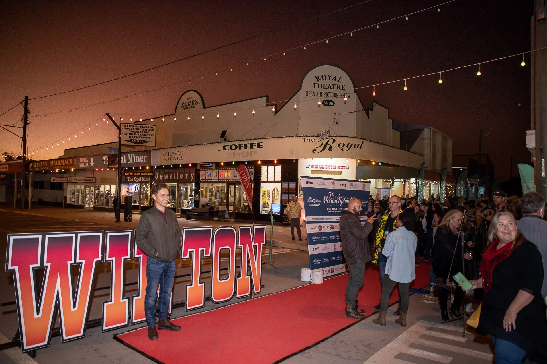 People gathered outside the Royal Theatre at night for an event, with some standing in line and others posing near a large 'WINTON' sign.