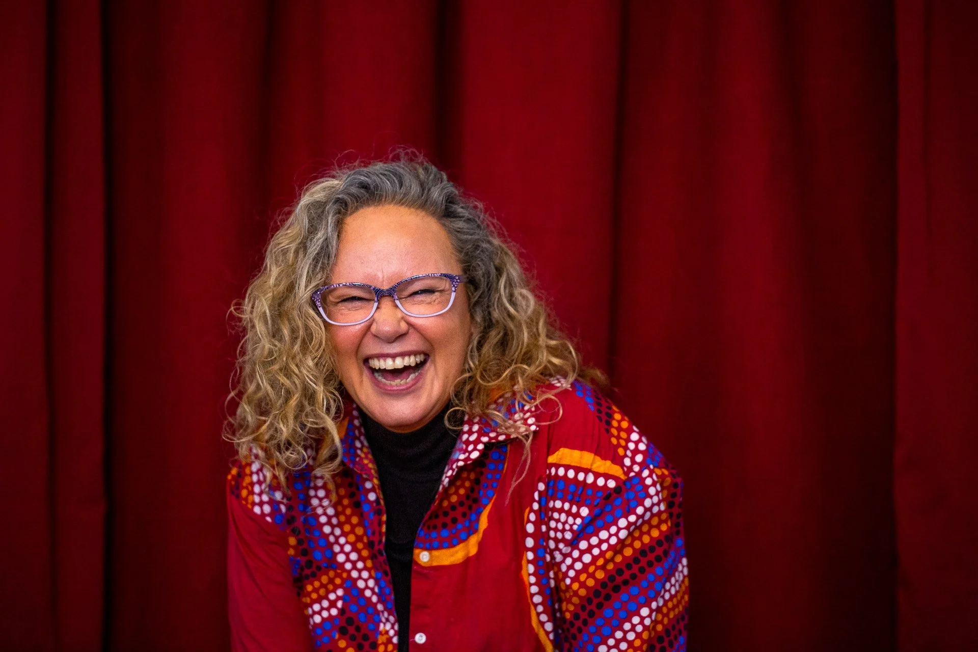 A woman with curly blonde hair wearing glasses and a colorful red, blue, and yellow patterned jacket, smiling in front of a red curtain.