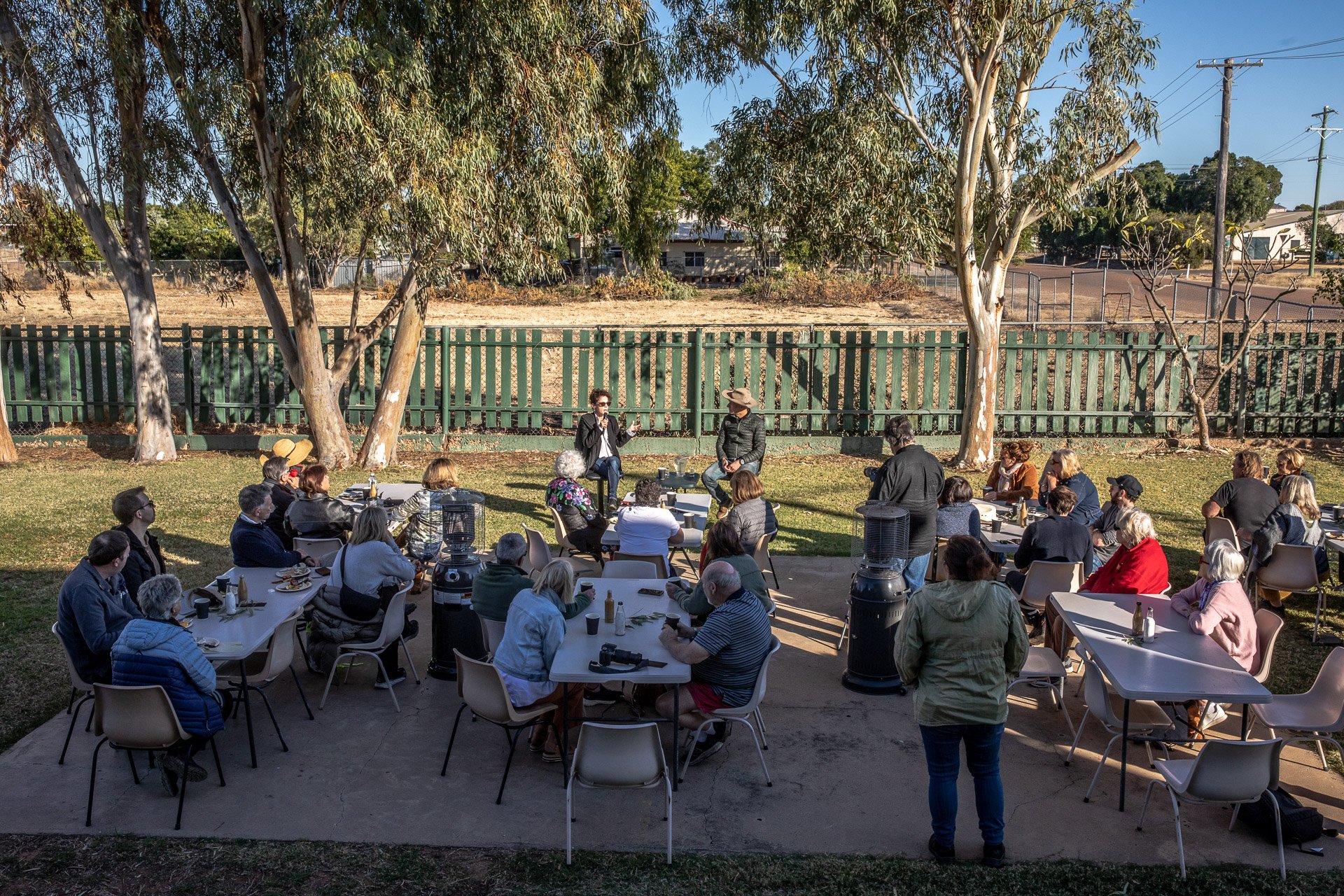 An outdoor gathering with people seated at tables, eating breakfast, listening to two speakers on a small stage in a yard with trees and a fence, under a clear blue sky.