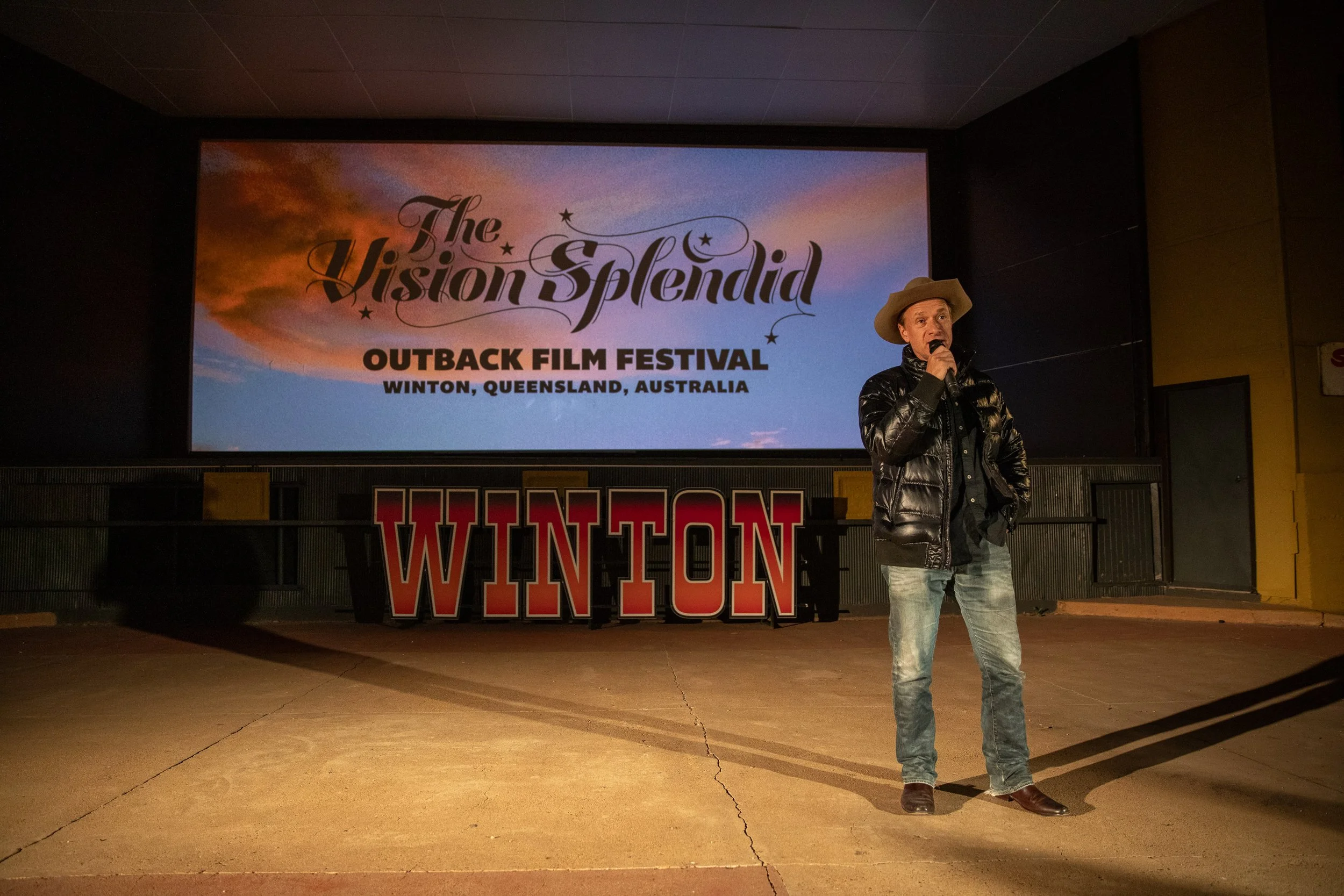 A person speaking into a microphone on stage with a large screen behind displaying the logo for The Vision Splendid Outback Film Festival in Winton, Queensland, Australia. The person is dressed in a cowboy hat, black puffer jacket, jeans, and brown b