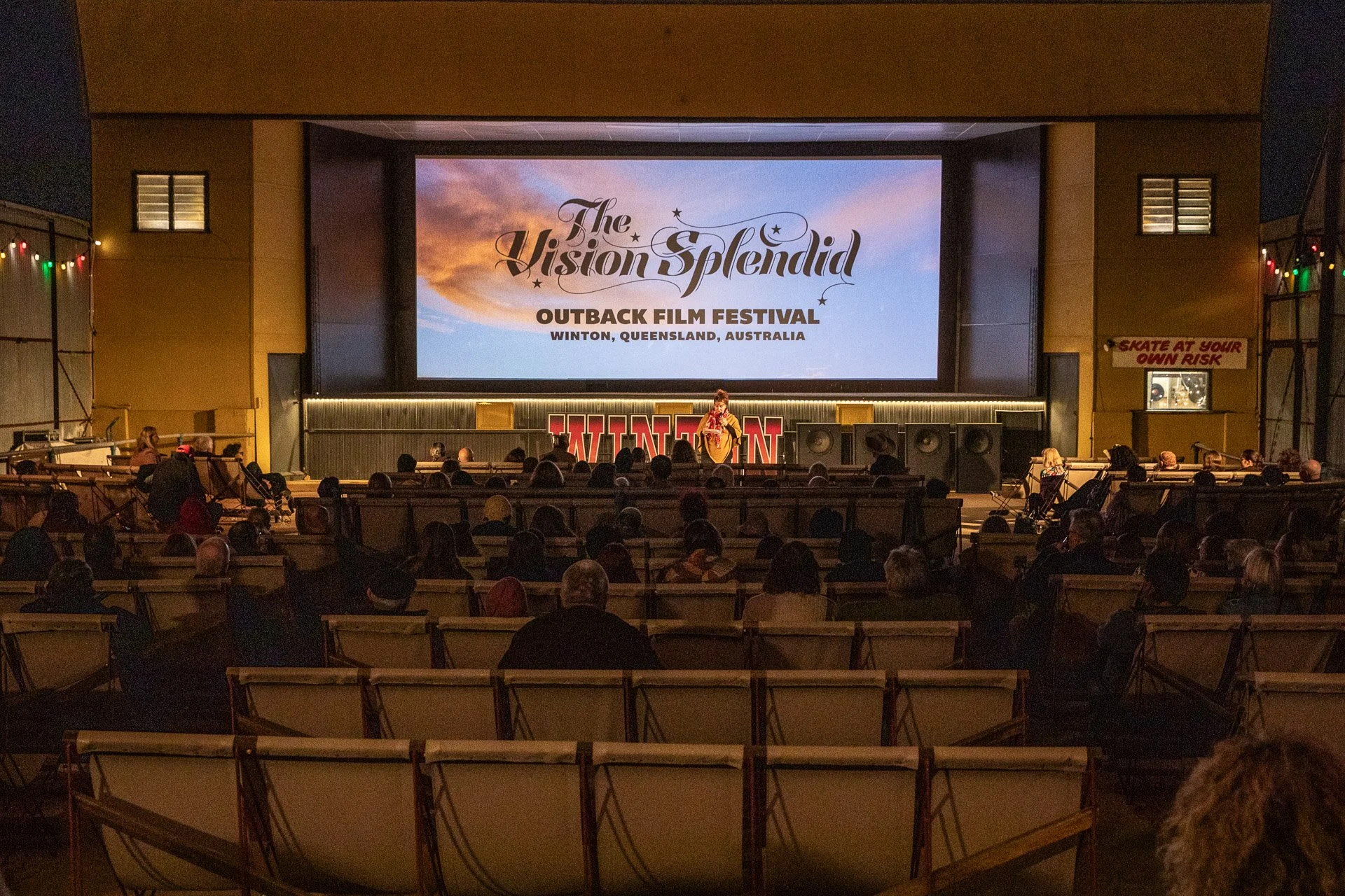 Audience watching a film screening at Outback Film Festival in Winton, Queensland, Australia. The scene shows people seated in an outdoor theatre with a large screen displaying the event's logo and location. A speaker is on stage near the screen.