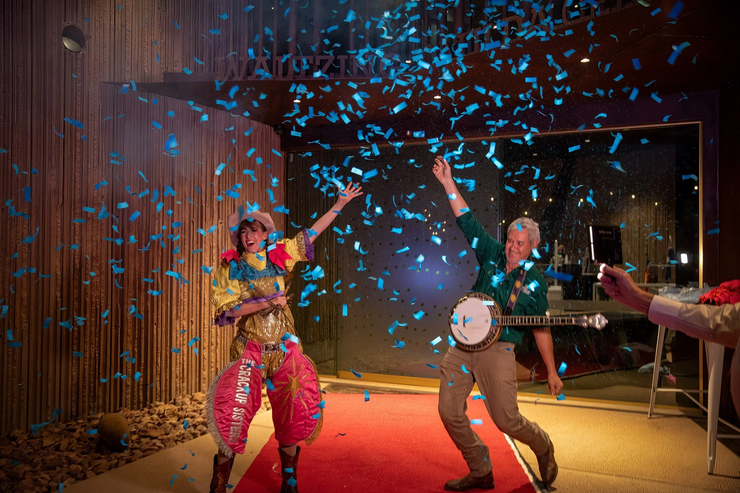 Two performers celebrating with blue confetti outside a concrete building. One is a woman dressed as a clown with colorful costume and a cowboy hat, and the other is a man playing a banjo, both smiling and enjoying the moment.