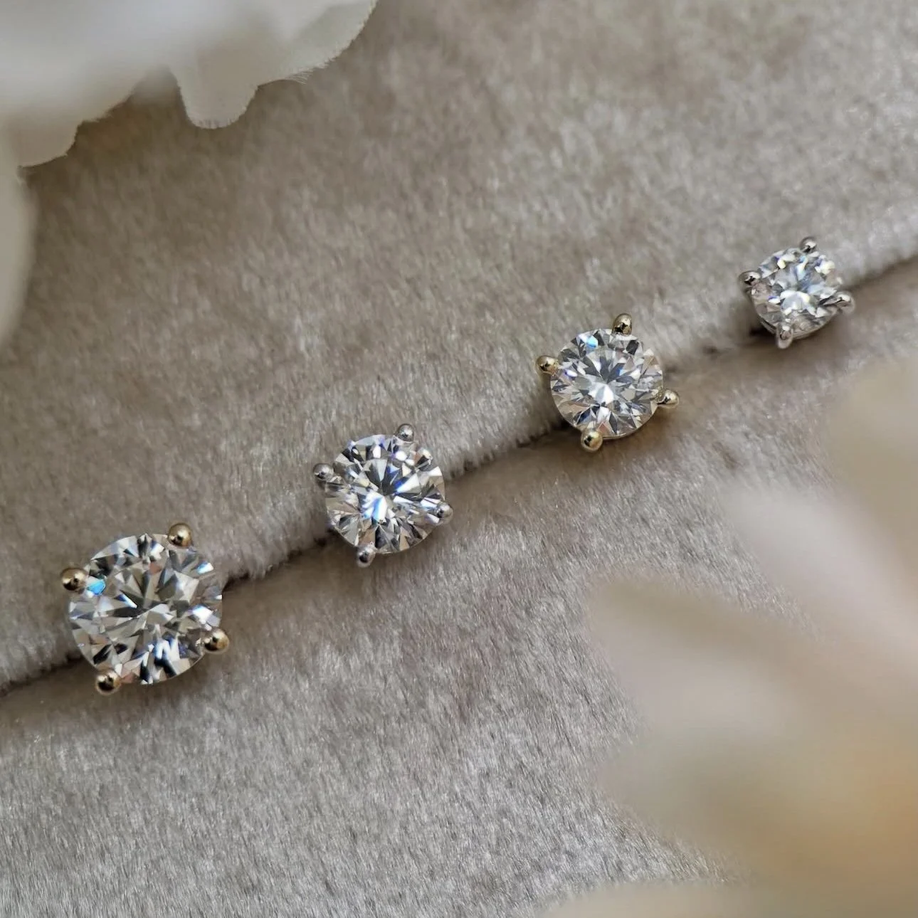 Four round cut diamond stud earrings, three with yellow gold prongs and one with silver prongs, displayed on a soft beige fabric background.