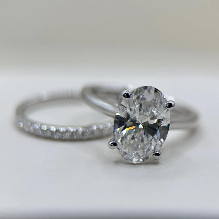 Close-up of a diamond engagement ring with a large oval-cut diamond and a wedding band with small diamonds, on a white surface.