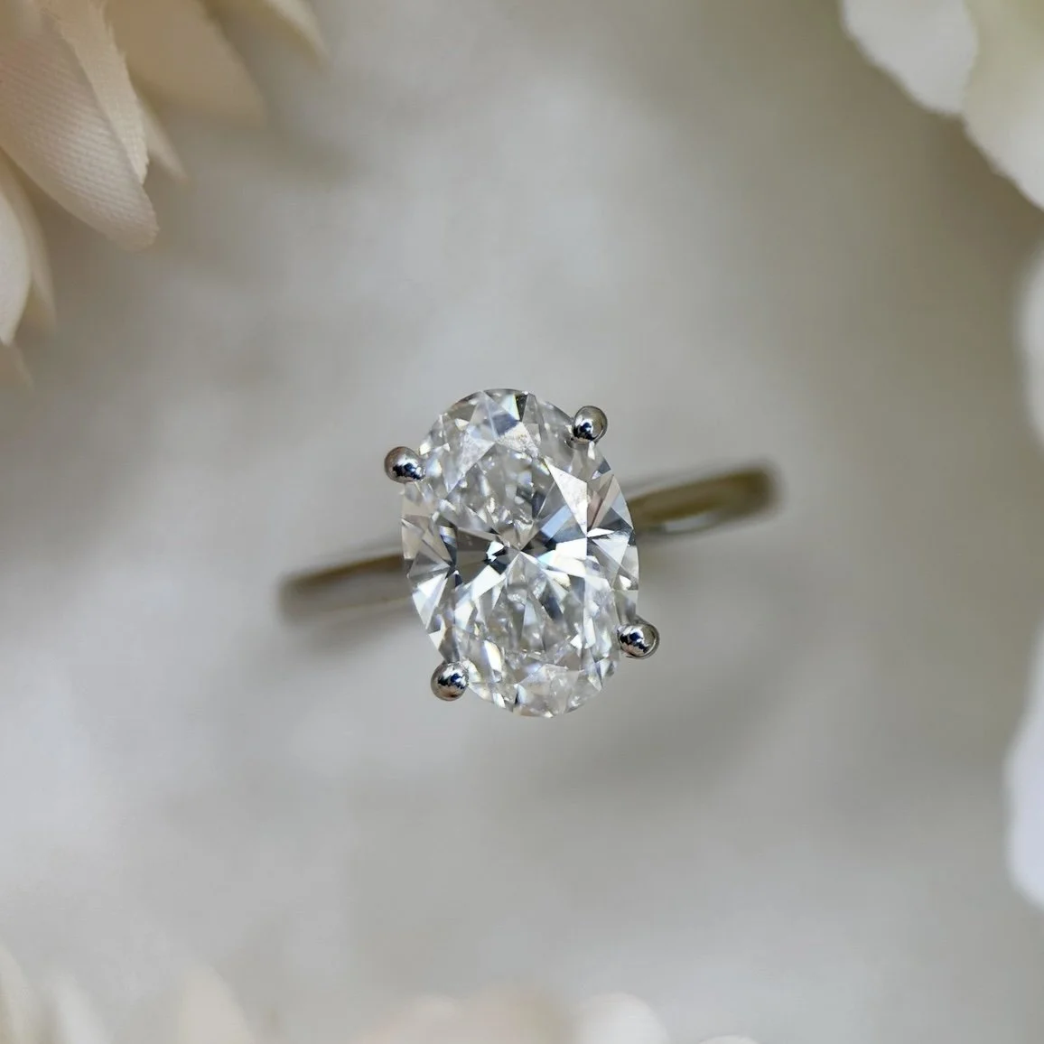 A close-up of an oval-shaped diamond engagement ring set in a silver band, surrounded by white flowers.