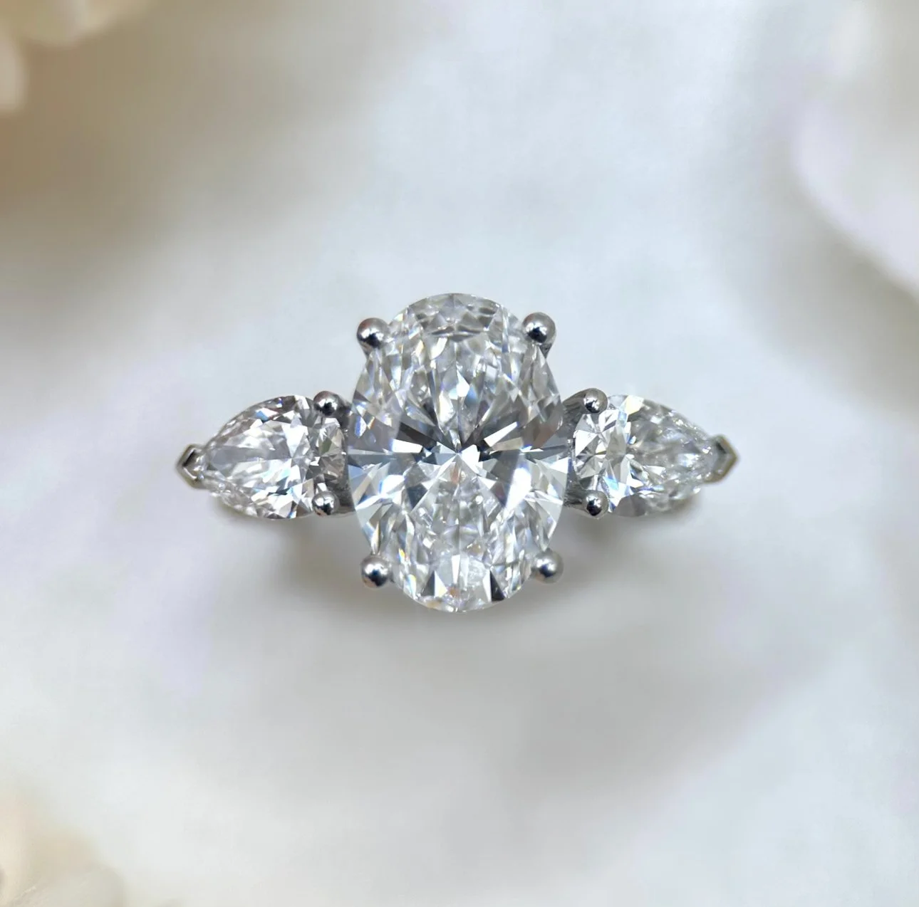 Close-up of a diamond ring with a large central oval diamond and four smaller side diamonds, set in a silver band, on a soft white background.