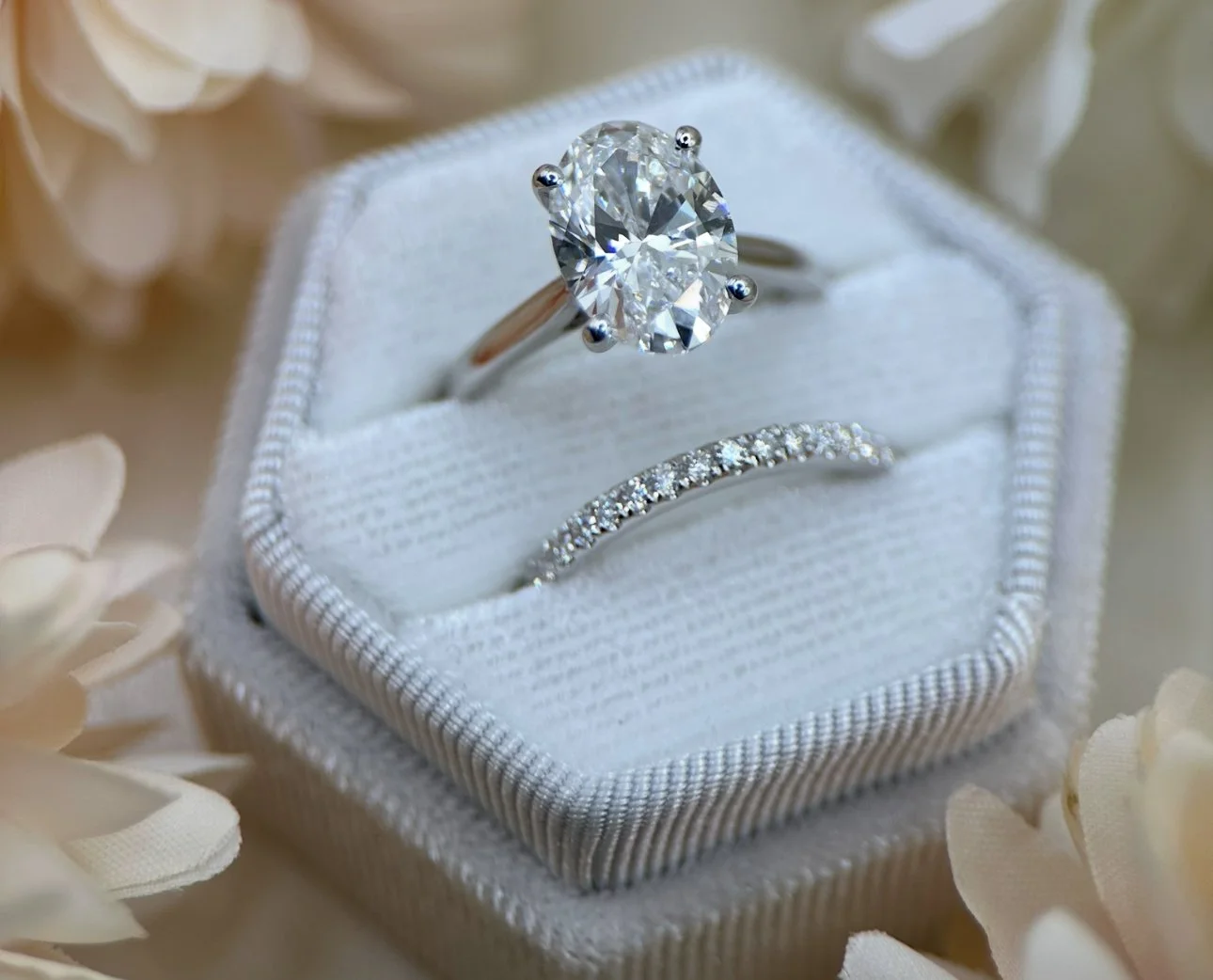 A diamond engagement ring with an oval center stone and a diamond wedding band in a white padded jewellery box surrounded by cream-colored flowers.