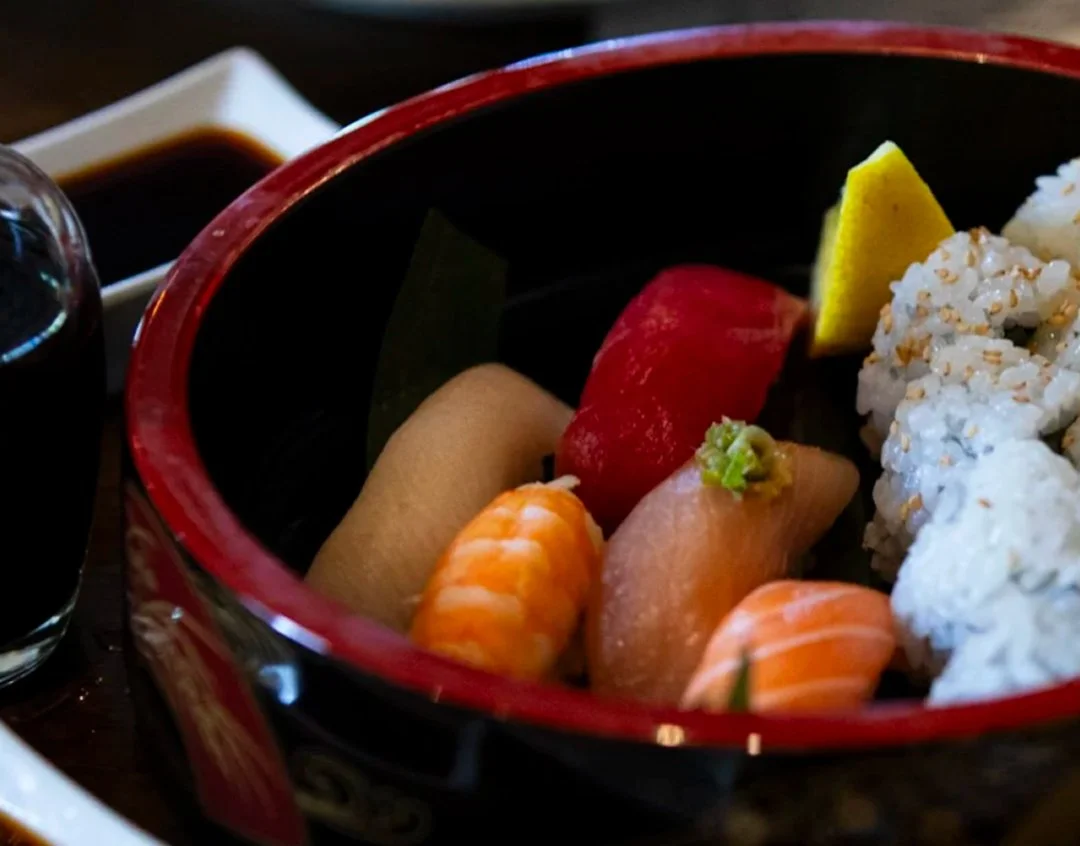 Assorted sushi pieces in a black and red bowl with wasabi and lemon wedge, accompanied by soy sauce and drink