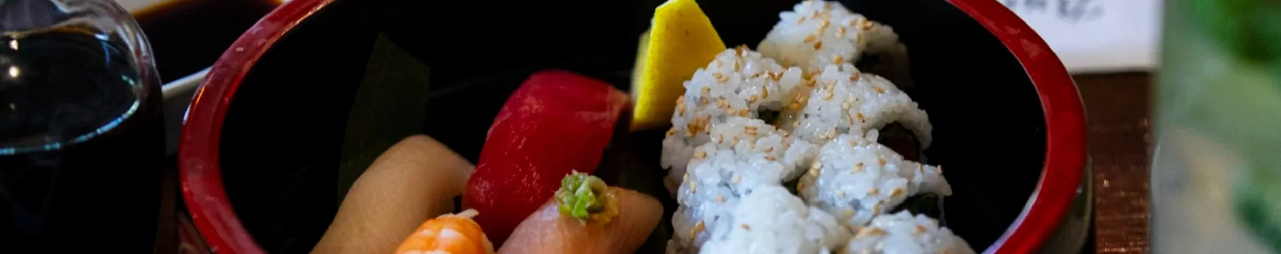 Close-up of a Japanese sushi platter featuring sliced sashimi, sushi rolls with sesame seeds, wasabi, and pickled ginger in a black and red bowl.