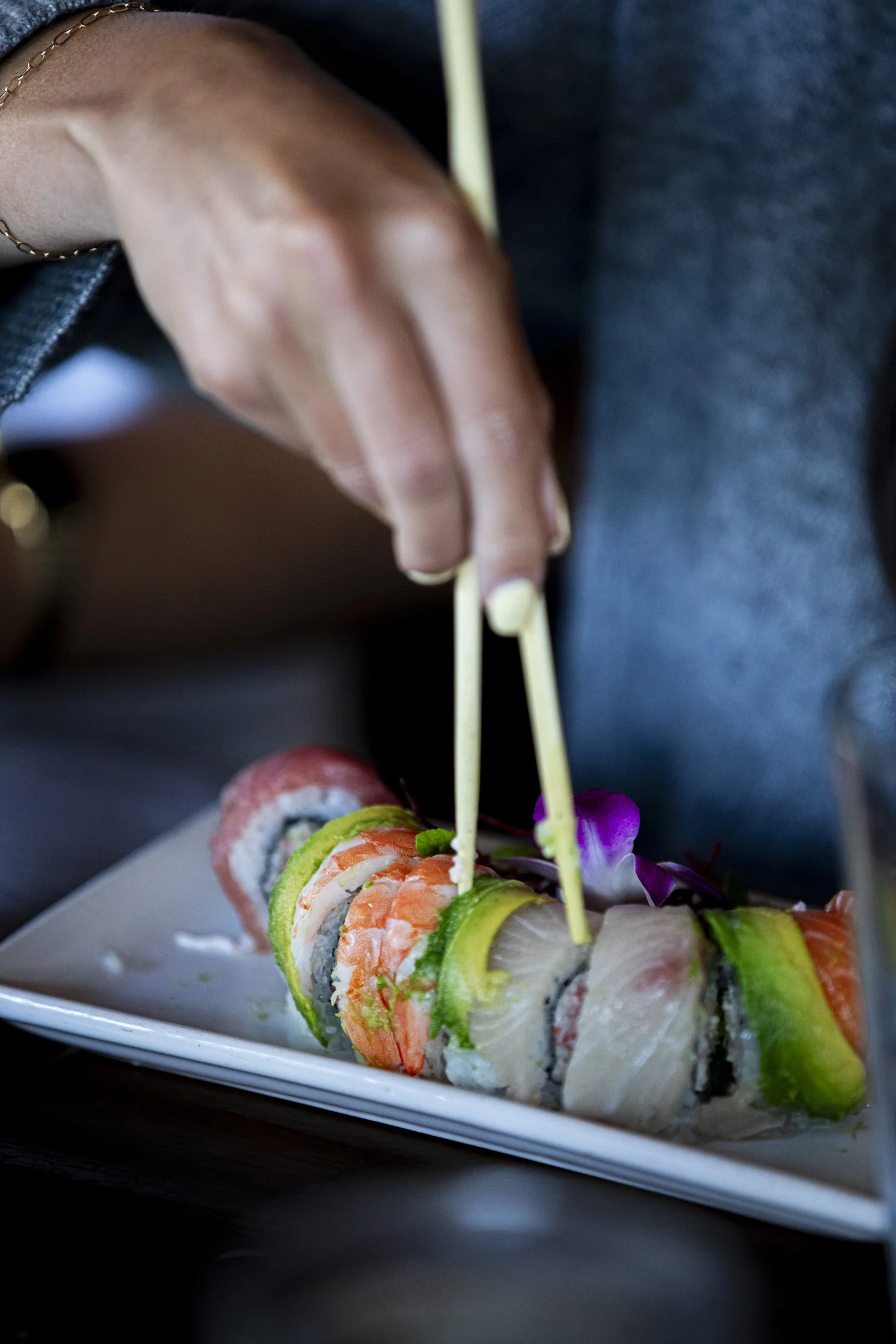 Close-up of a person's hand using chopsticks to pick up a piece of sushi on a white rectangular plate, with various colorful sushi rolls decorated with edible flowers.
