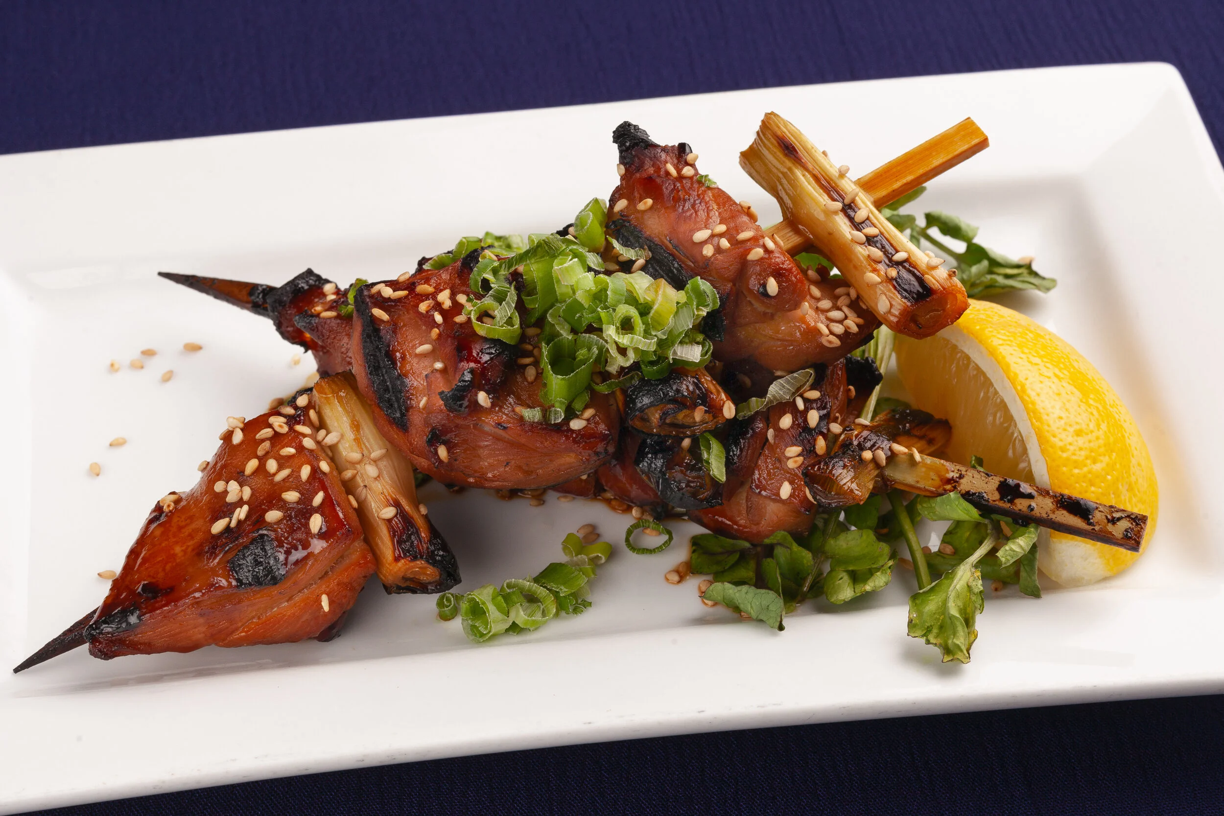 Grilled chicken skewers with green onions and sesame seeds, served with lemon wedge and greens on a white plate.