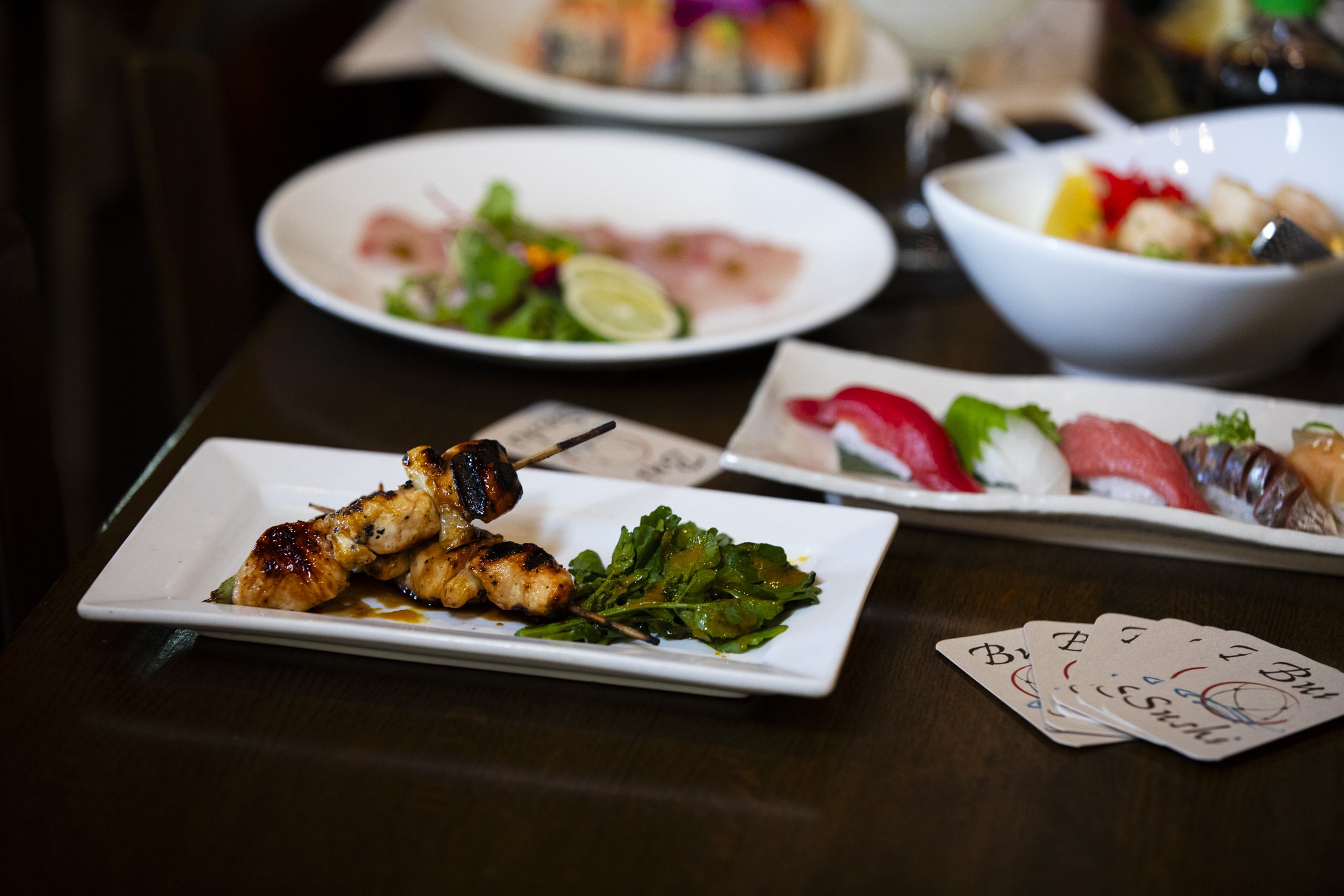 Assorted Japanese dishes including grilled chicken skewers, sushi, sashimi, and vegetable dishes on a dark wooden table.