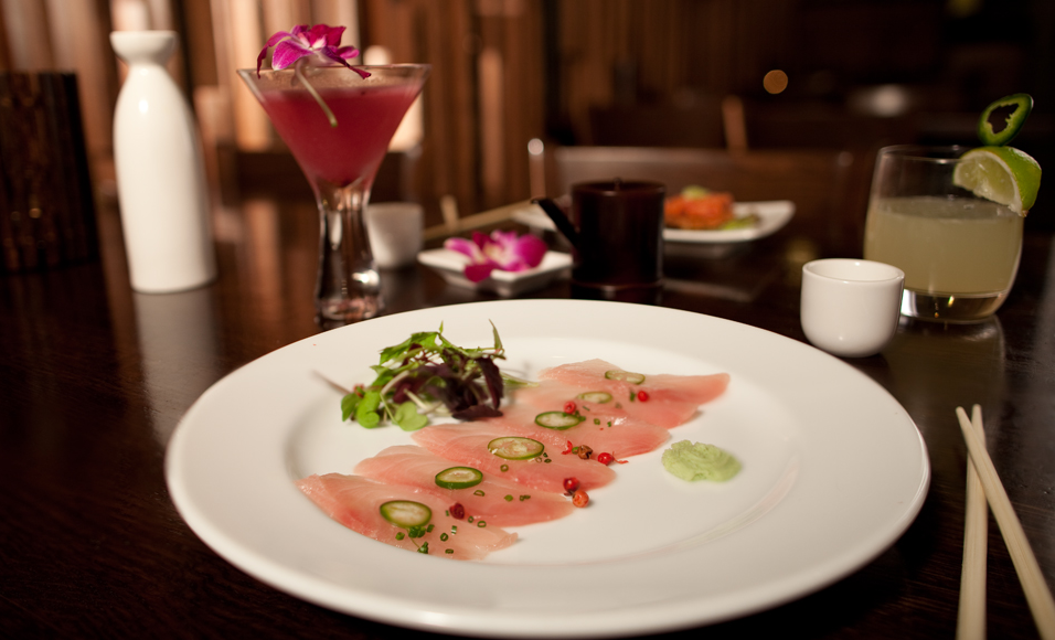 Plate of sashimi with sliced raw fish garnished with sliced peppers and green wasabi, accompanied by cocktails on a dark restaurant table.