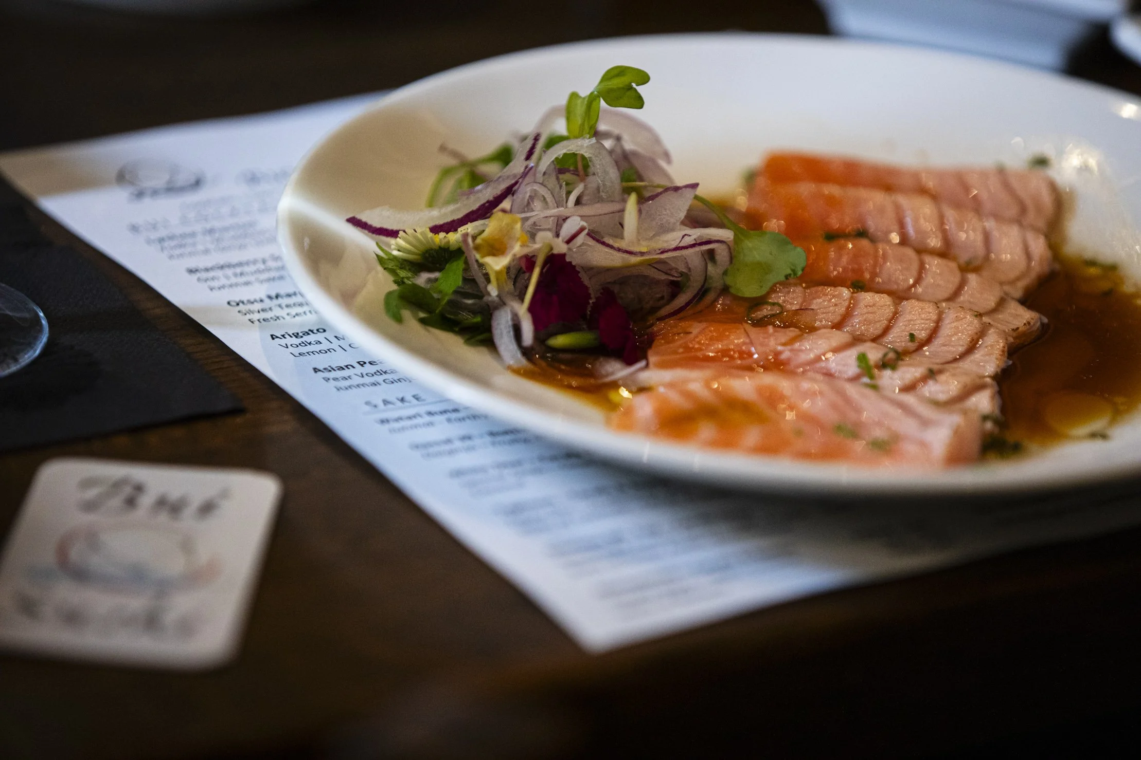 Sliced raw salmon fish served on a white plate with a side of mixed greens and salad. The plate is placed on top of a menu on a wooden table.
