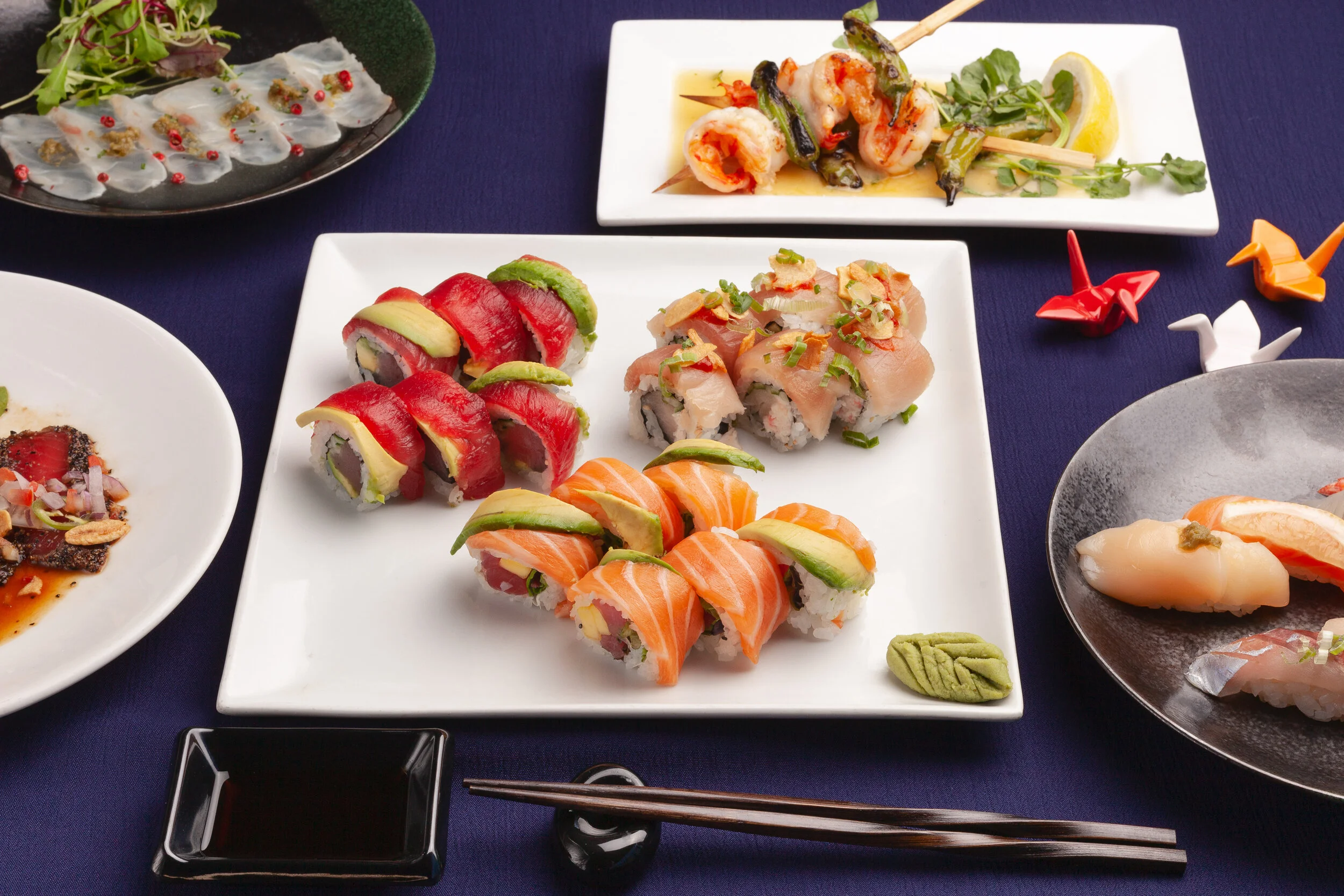 Assorted sushi rolls on white and black plates, with fresh sashimi, seafood skewers, wasabi, and decorative origami on a navy blue tablecloth.