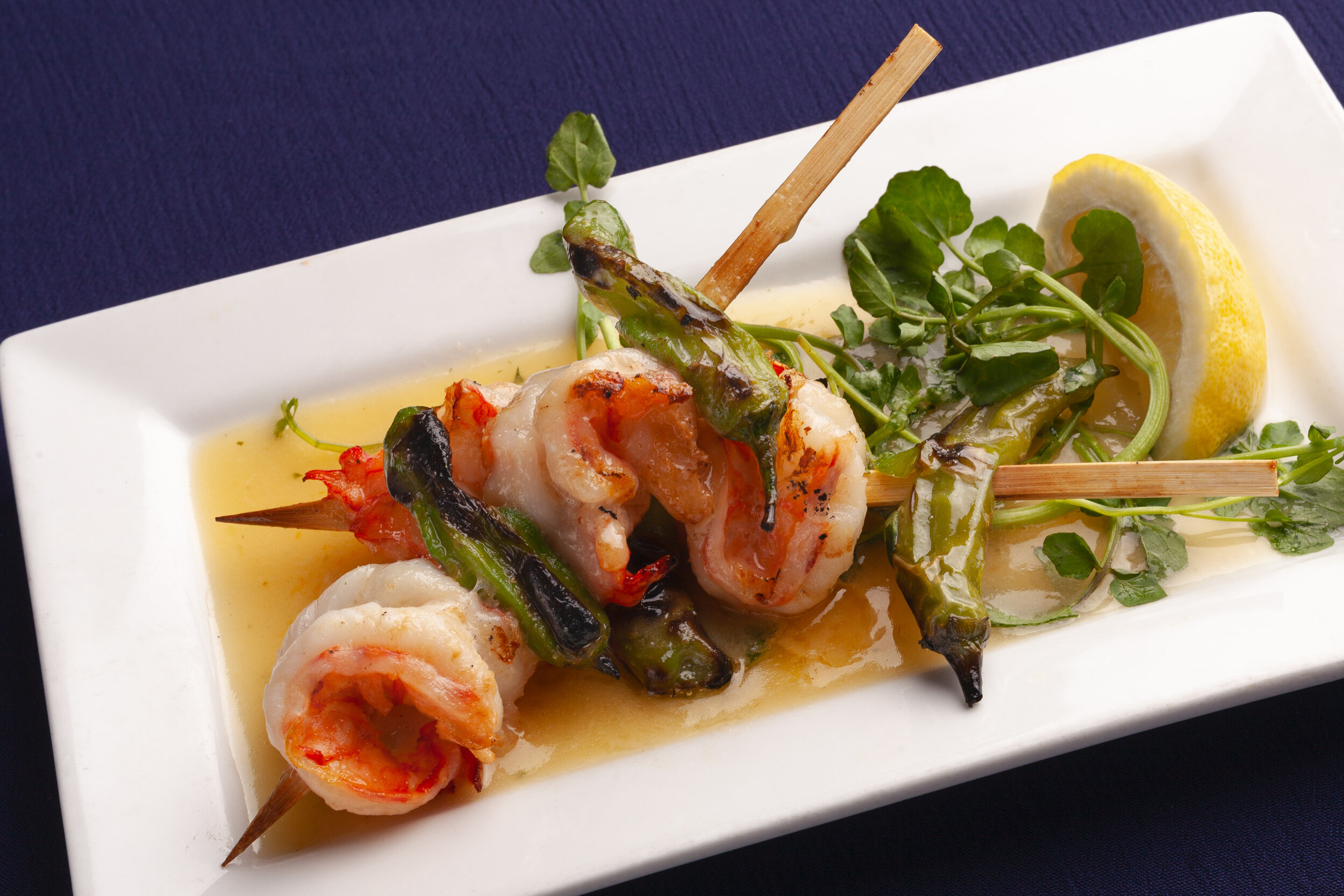 Shrimp and vegetable skewers served on a white rectangular plate with lemon wedge and greenery garnishes.