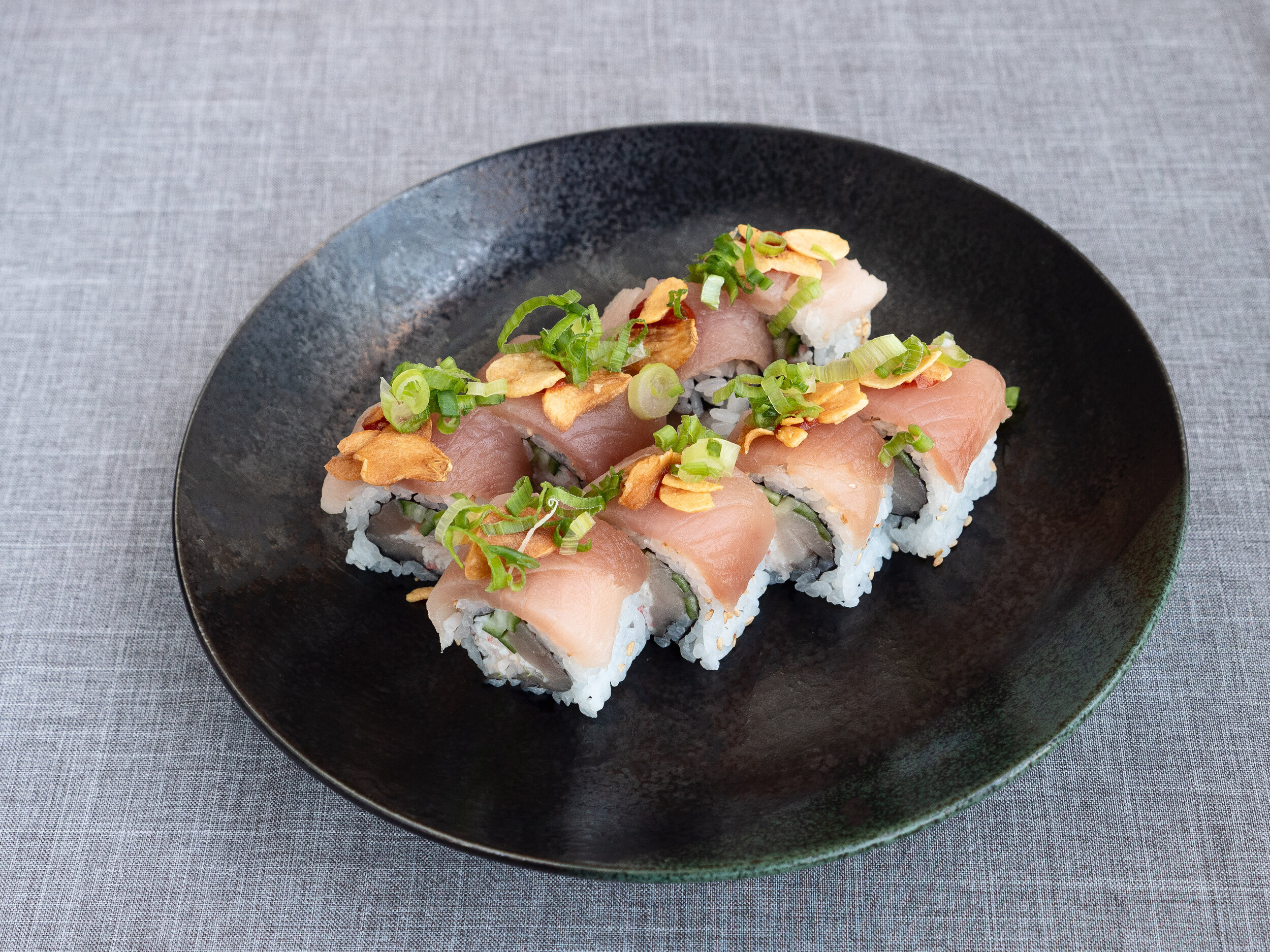 Sushi rolls topped with sliced garlic, chopped green onions, and garnished with fish slices on a black plate.