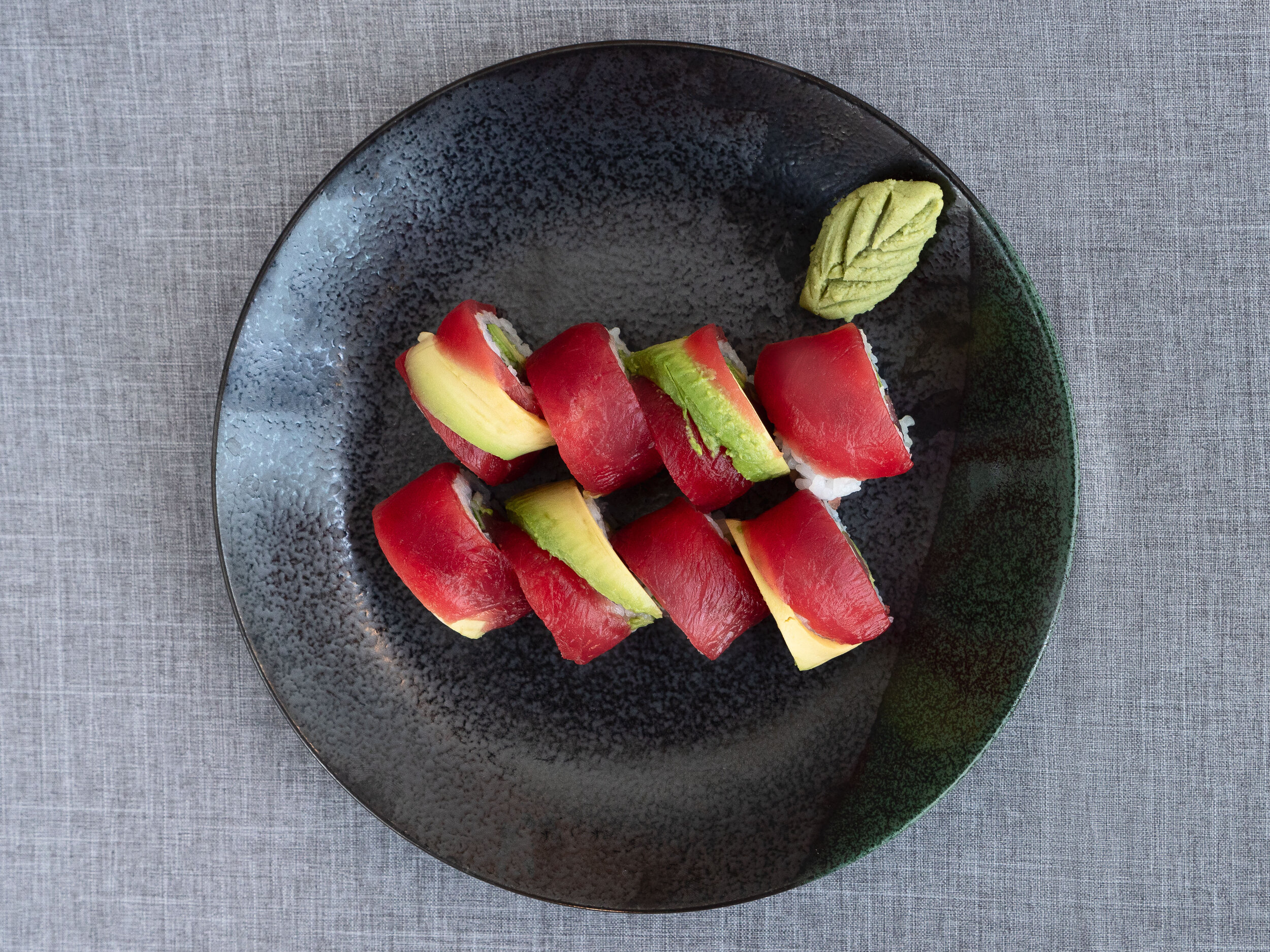 Sushi rolls with tuna and avocado on a black plate, accompanied by wasabi, placed on a gray tablecloth.