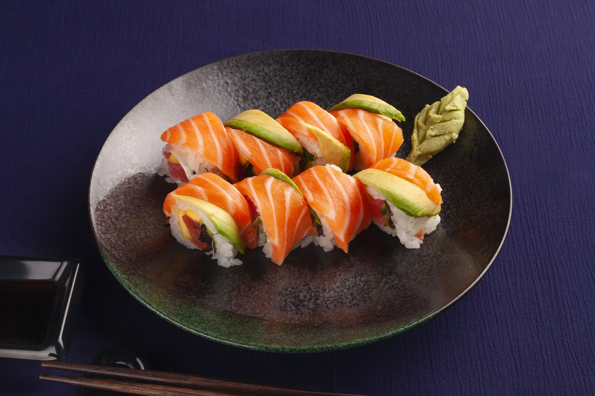Sushi rolls topped with sliced salmon, avocado, and cream cheese, served with a wasabi dollop on a black plate.